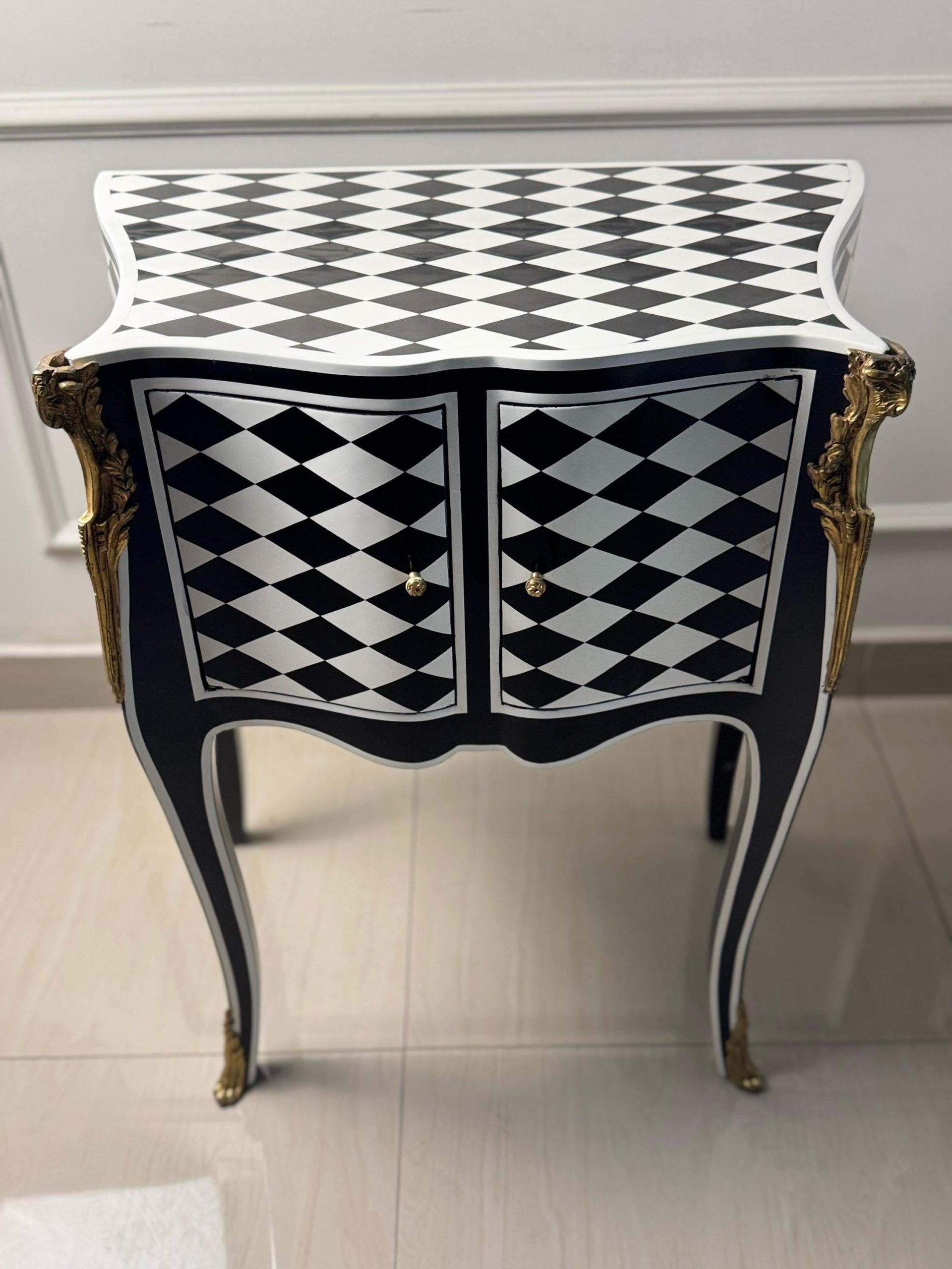 May include: A black and white checkered end table with gold accents and two doors. The table has a curved top and legs.