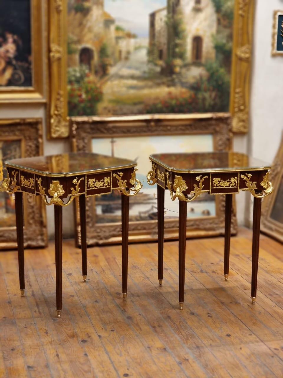 May include: Two antique side tables with dark wood frames, glass tops, and ornate gold-colored accents. The tables have four slender legs with gold-colored tips. The tables are displayed in a room with framed paintings.