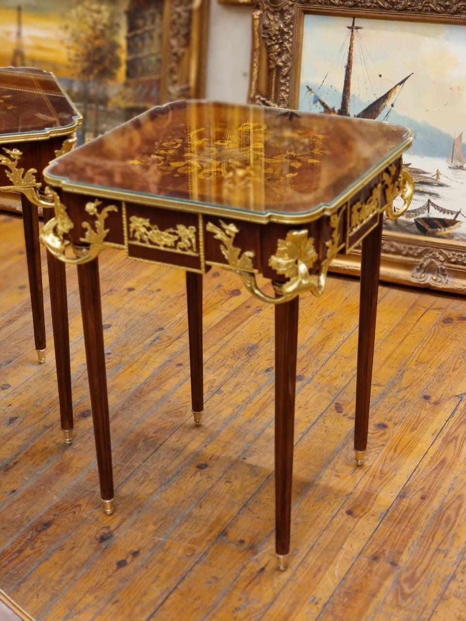 May include: A pair of antique side tables with a square top and four slender legs. The tables feature a dark wood finish with gold-colored accents, including ornate detailing around the edges and legs. The tabletop has a reflective surface.