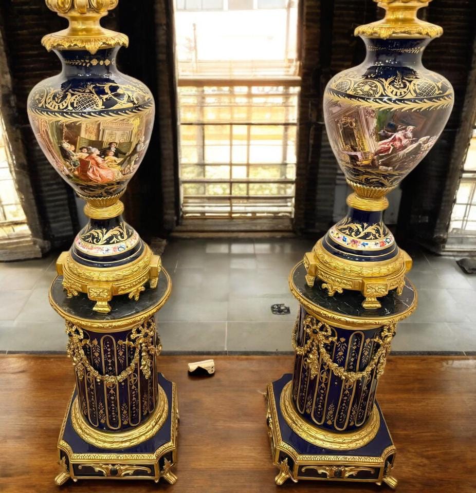 May include: Two ornate, antique vases with a dark blue and gold color scheme. The vases feature detailed painted scenes and are mounted on decorative pedestals. The pedestals have a dark blue base with gold accents and ornate detailing.