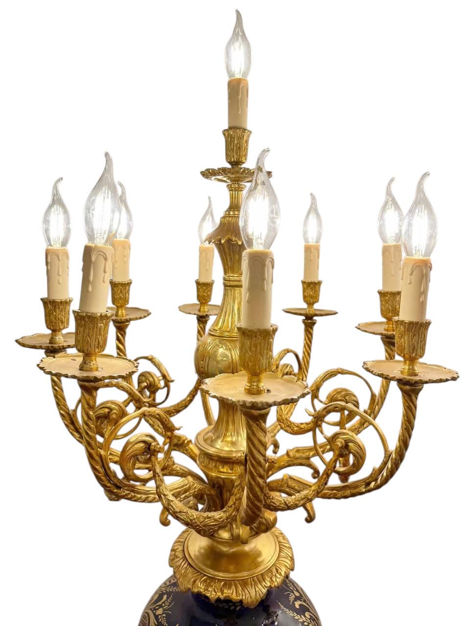 May include: A gold-toned candelabra with ten faux candle lights. The base is a dark blue with a floral pattern. The candelabra has ornate, swirling arms and a central column. The candle lights have a flame-shaped design.