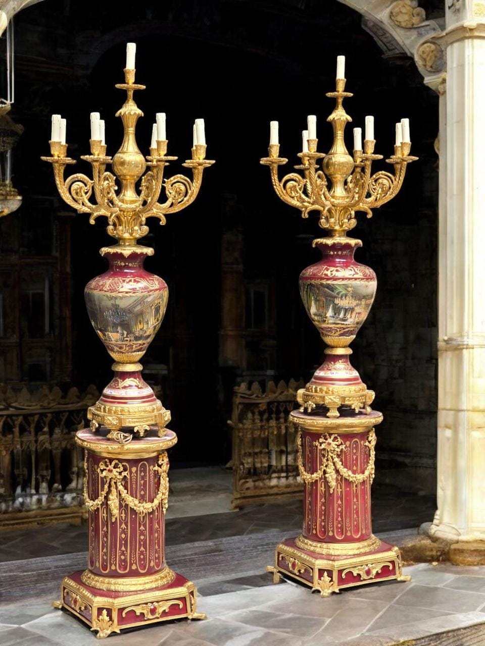 May include: Two ornate, gold-colored candelabras with multiple candle holders. The candelabras have a red and gold base with painted scenes. The design includes intricate details and a classical aesthetic, suitable for home decor.