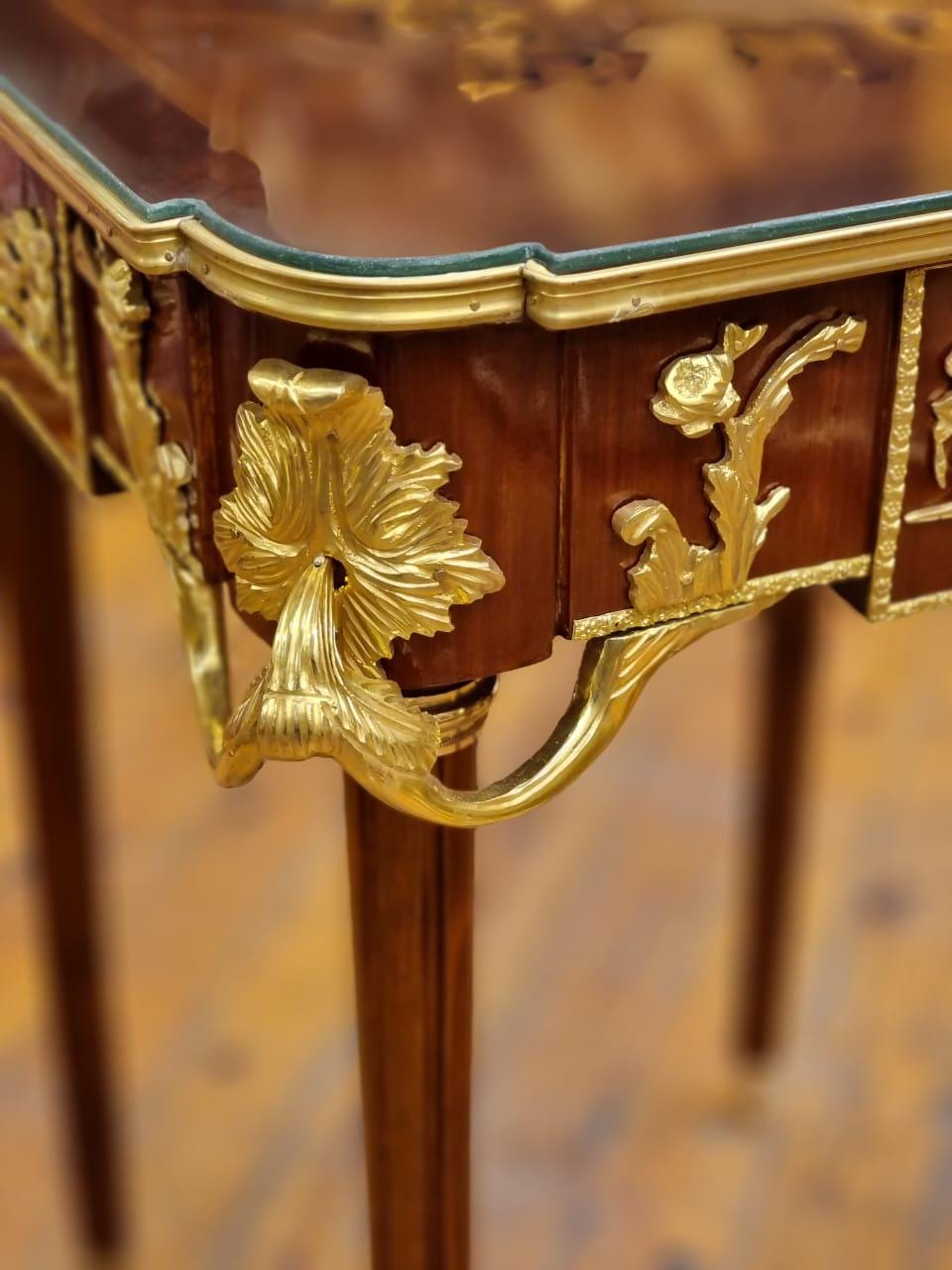 May include: Close-up of a detailed antique table. The table features a dark wood finish with ornate gold-colored metal accents. The design includes floral and leaf motifs, with a glass top and slender legs. The table's craftsmanship is evident in the intricate details.