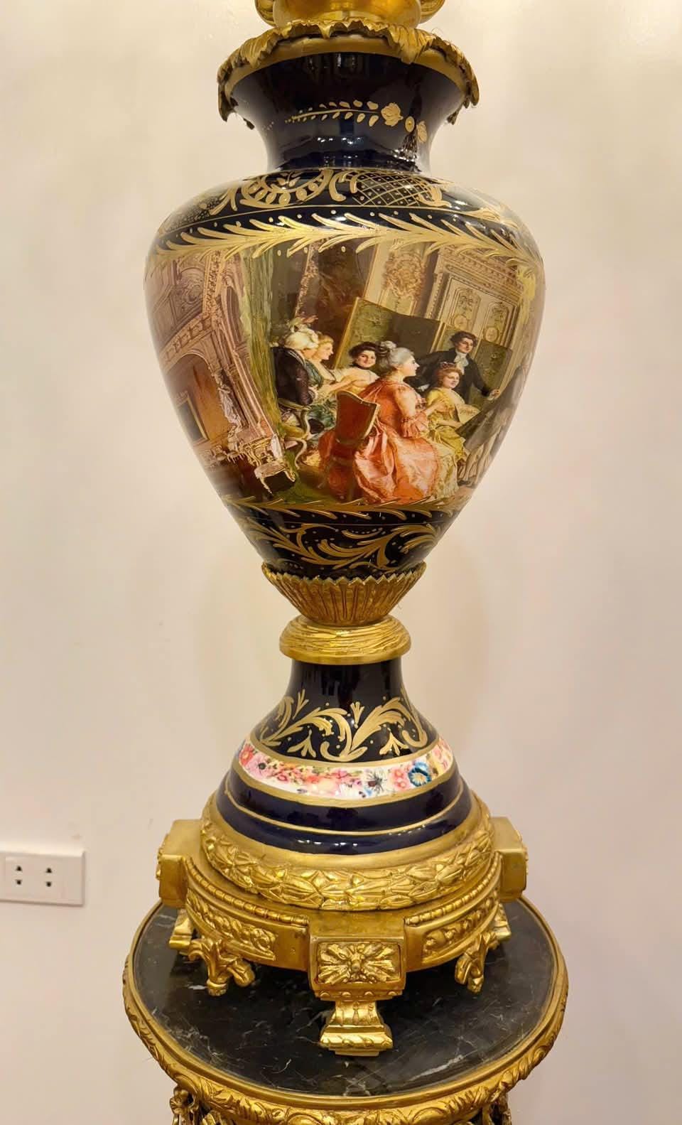 May include: A large, ornate vase with a dark blue base and gold accents. The vase features a painted scene of people in period clothing. The base has a gold, detailed design, and the vase sits on a round, black and gold stand.
