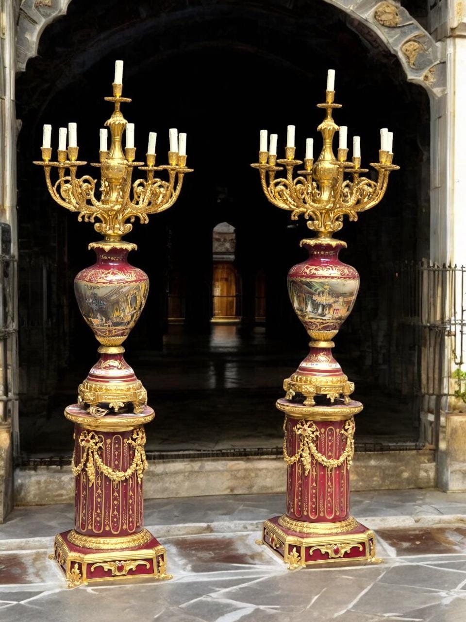 May include: Two ornate, antique candelabras with gold-colored arms and bases. The candelabras have a burgundy-colored vase-shaped center with a painted landscape scene. Each candelabra holds multiple white candles and stands approximately 6 feet tall.