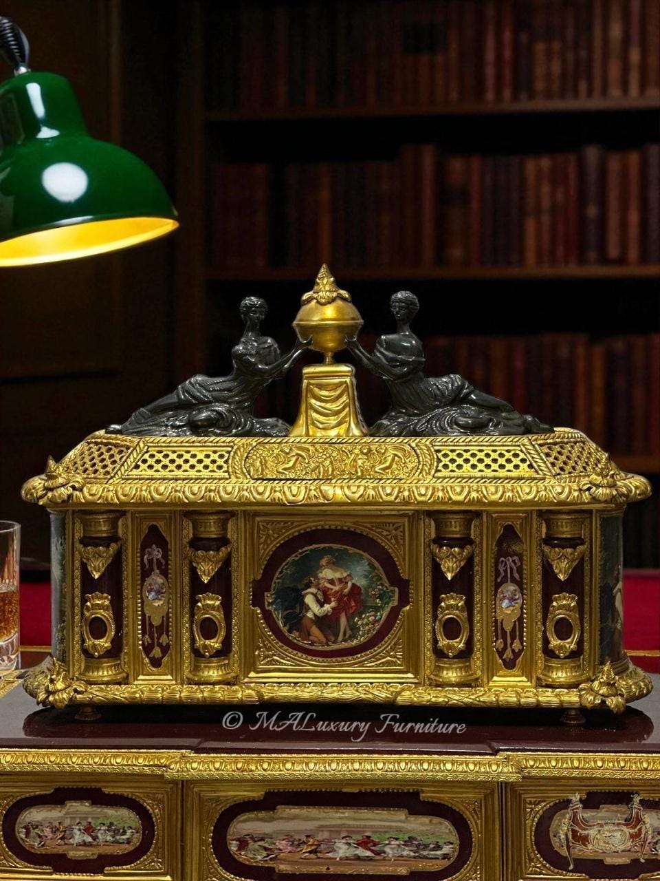 May include: Ornate gold-toned box with intricate detailing, featuring a central painting and decorative figures. The box has a burgundy background and is topped with two dark figures. A green lamp is visible in the upper left corner.