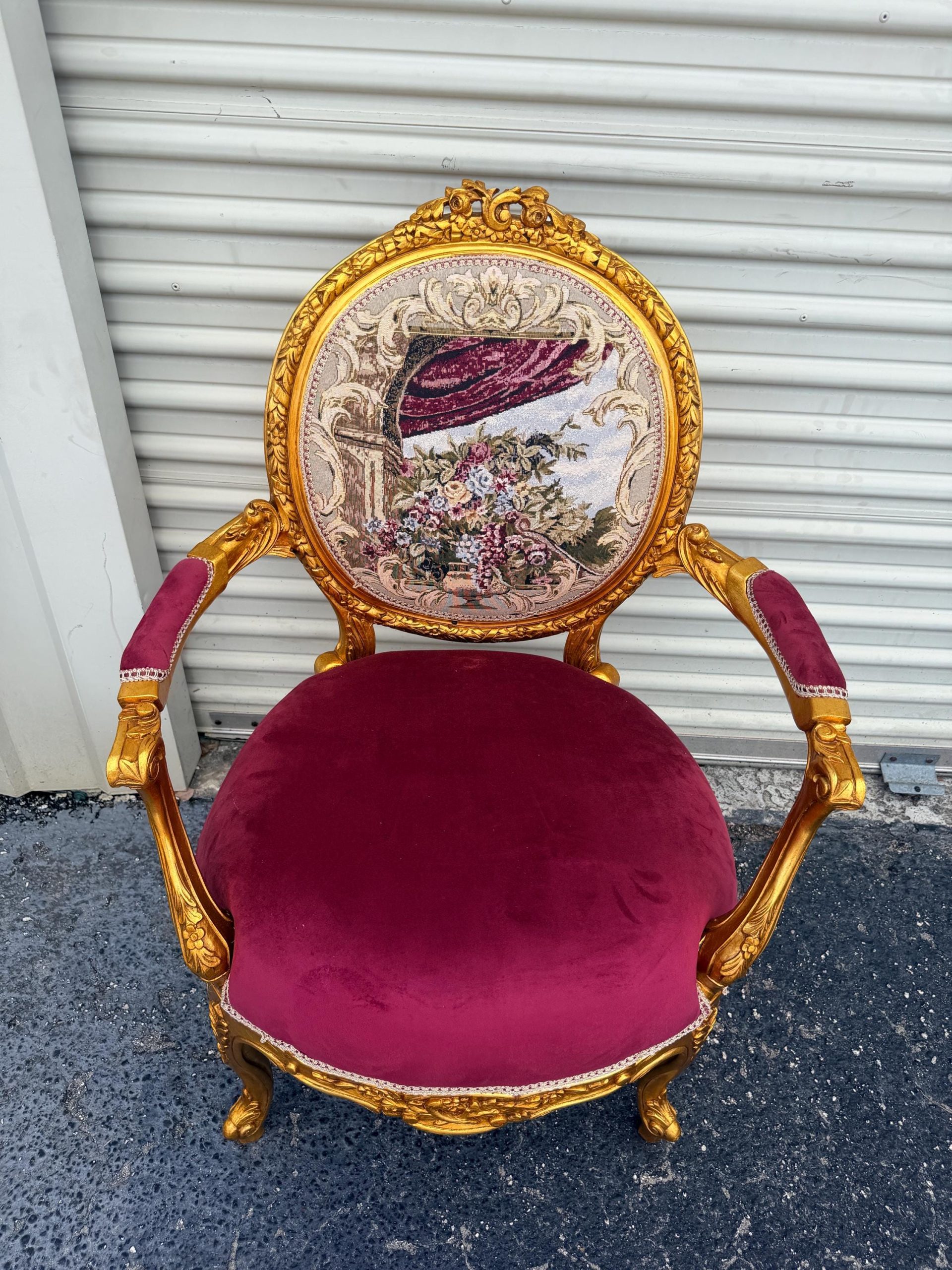 May include: Ornate antique chair with a gold-colored frame and burgundy velvet seat. The backrest features a tapestry with floral and curtain designs. The armrests are burgundy velvet. A decorative piece for a living room or bedroom.