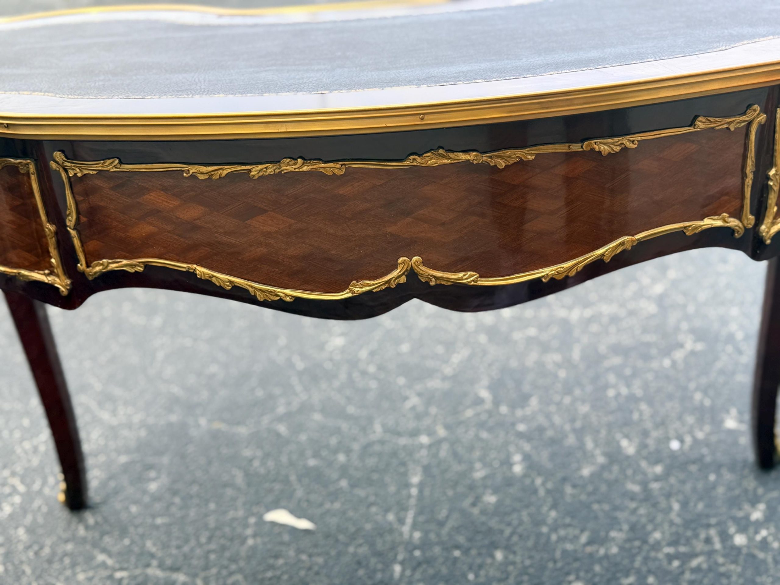 May include: Close-up of a mahogany-colored antique table with a black top and gold trim. The table features ornate gold detailing along the edges and a patterned wood grain design on the front panel. The legs are a dark mahogany color.