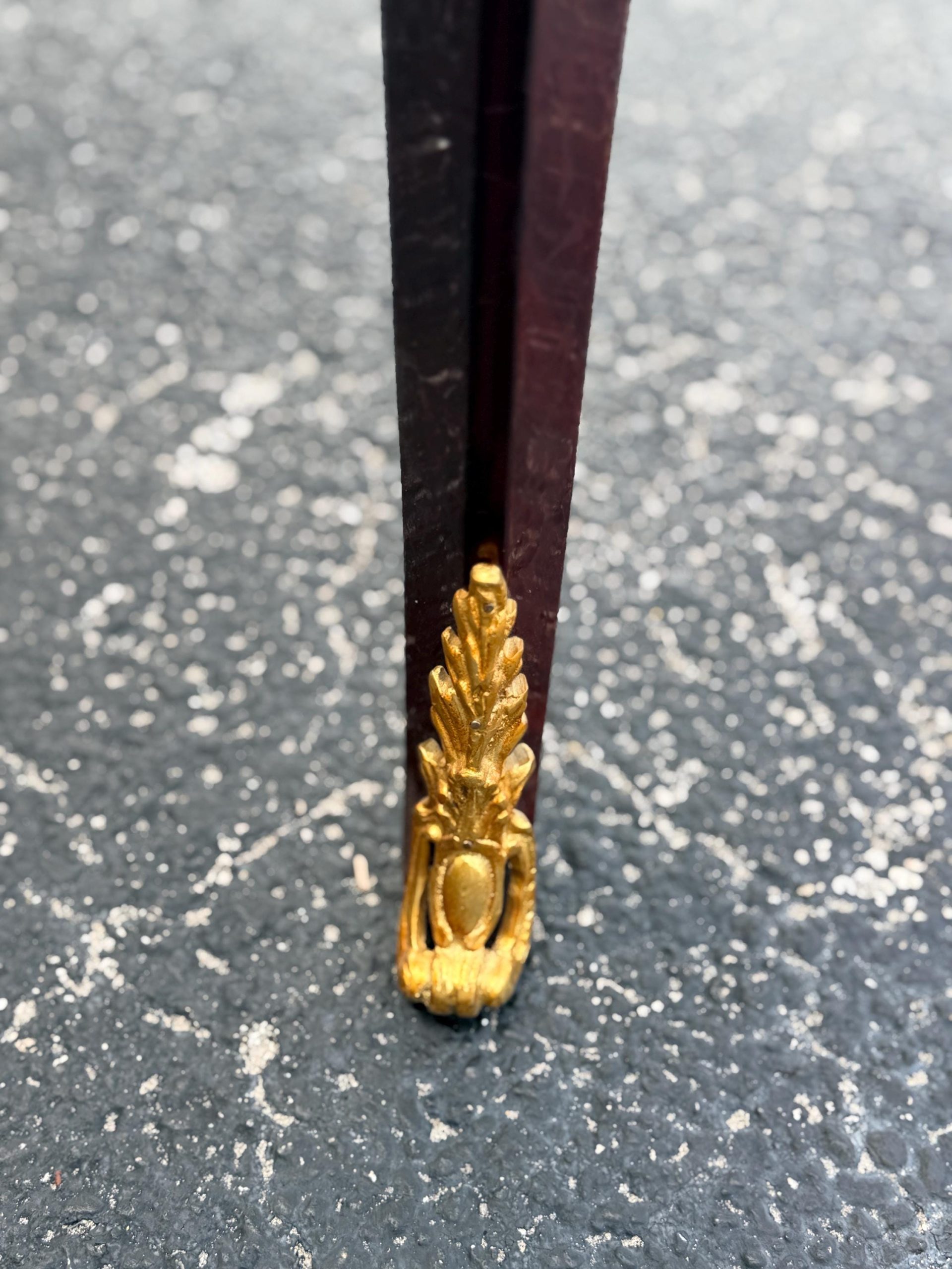 May include: Close-up of a dark brown table leg with a gold-colored decorative foot. The foot has a detailed, ornate design, possibly made of metal. The leg appears to be made of wood and has a rectangular shape.