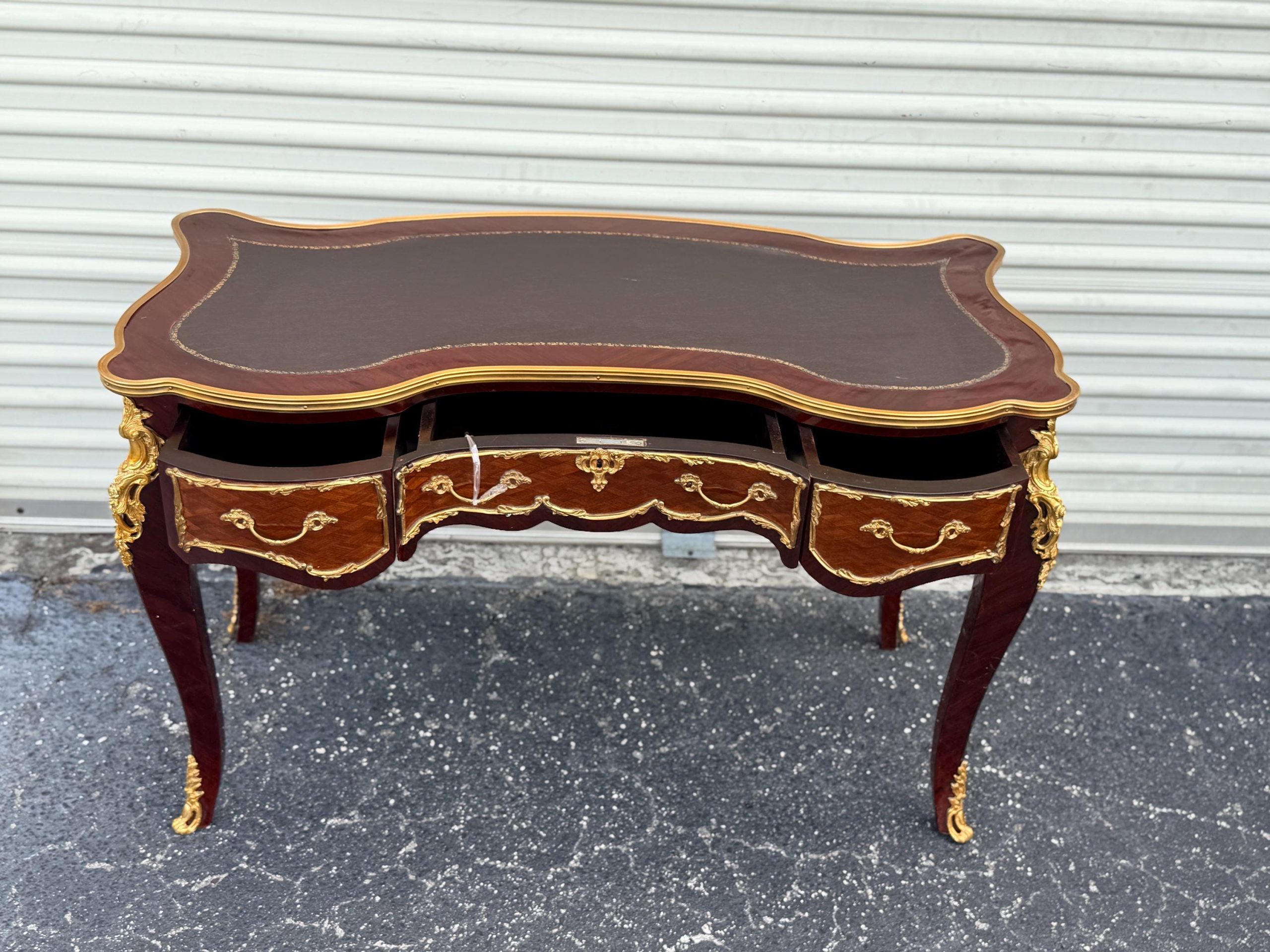 May include: A mahogany desk with a dark brown leather top and three drawers. The desk features ornate gold accents on the legs and drawer fronts. The desk has a curved design and is a classic piece of furniture.
