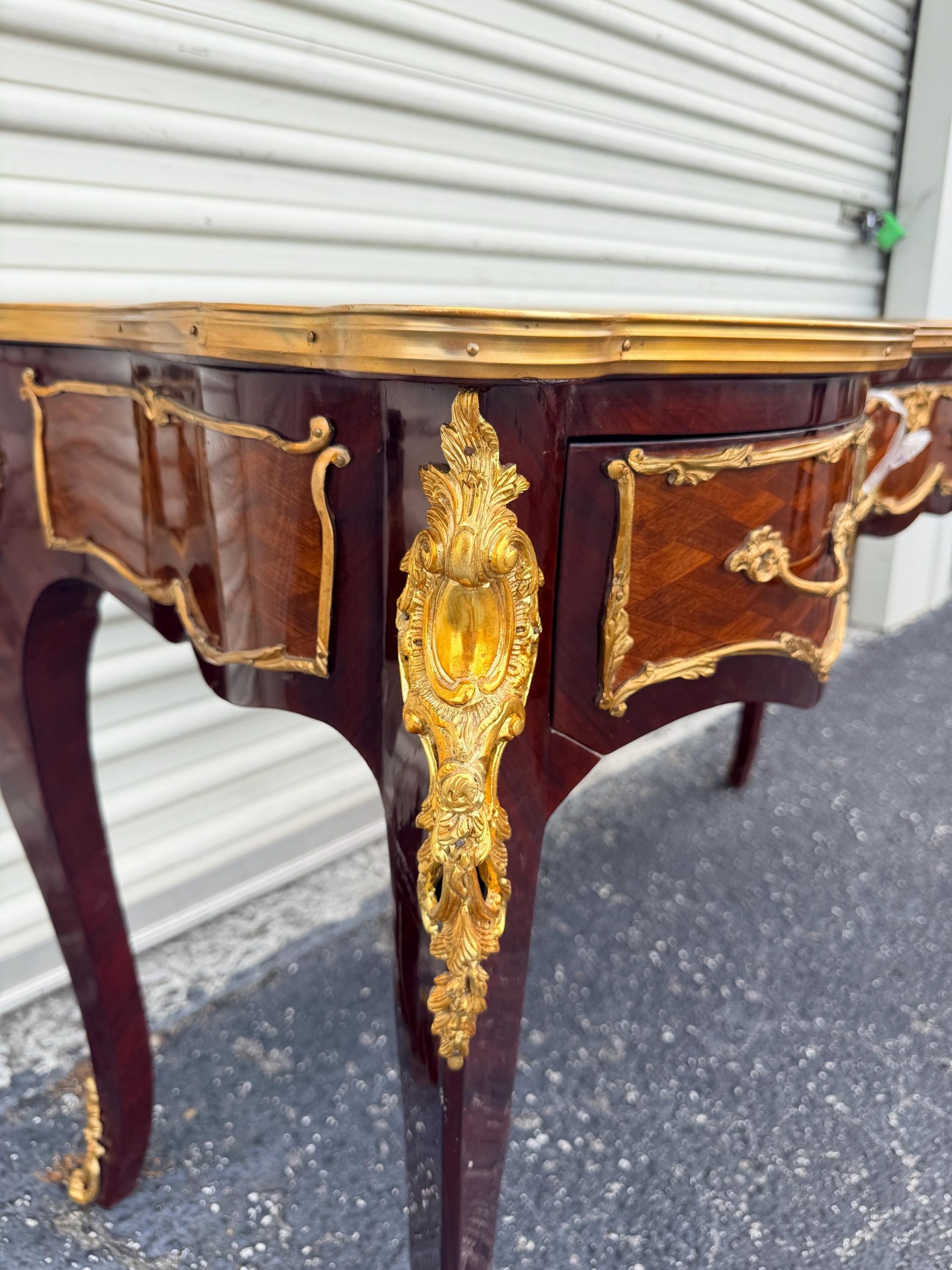 May include: A close-up of a mahogany desk with gold accents. The desk features a curved design with ornate gold detailing on the legs, drawer fronts, and edges. The wood grain is visible, and the desk has a glossy finish.