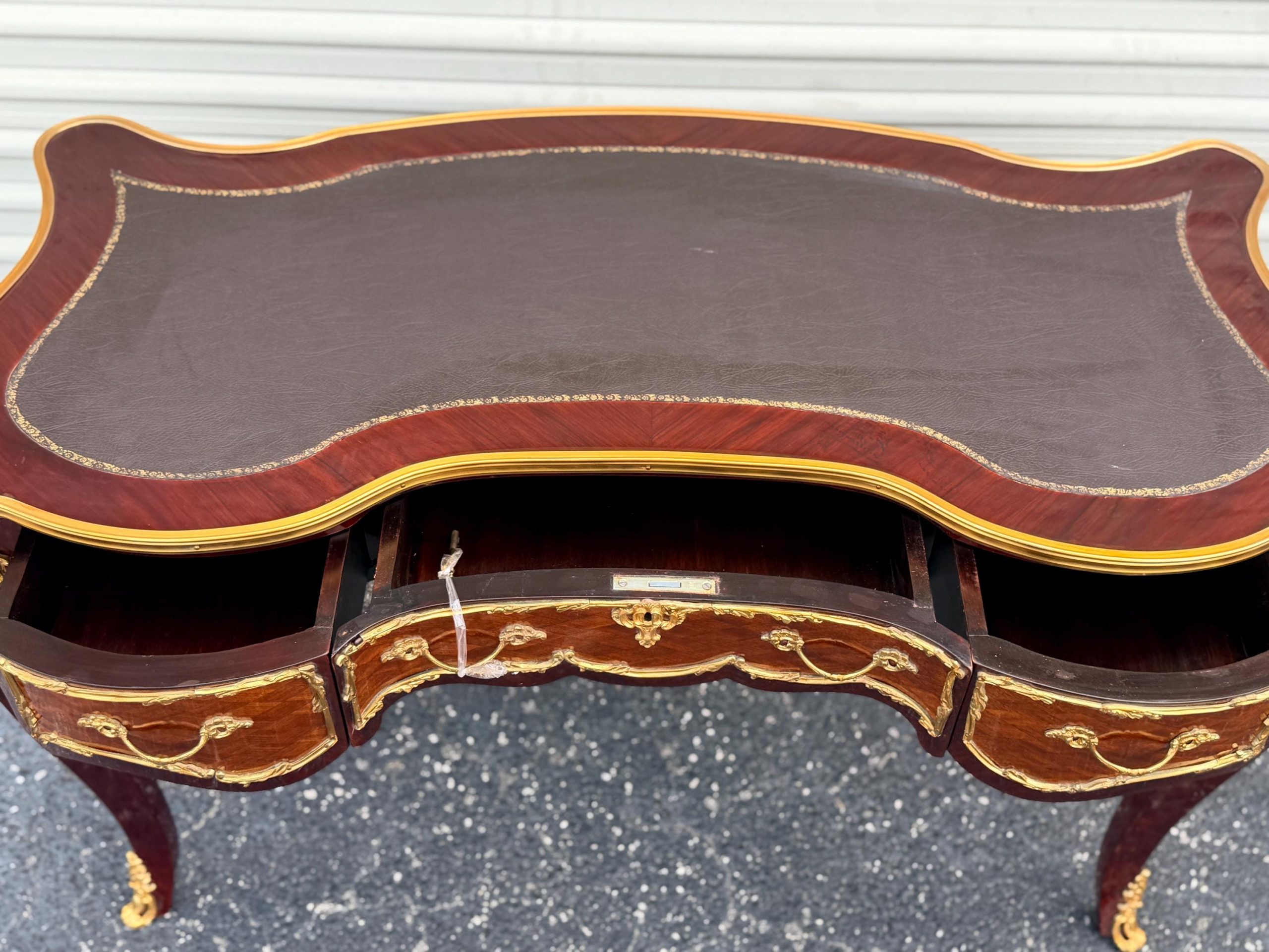 May include: A mahogany desk with a dark brown leather top and gold trim. The desk has three drawers with gold hardware and ornate legs. The desk is a classic piece of furniture.