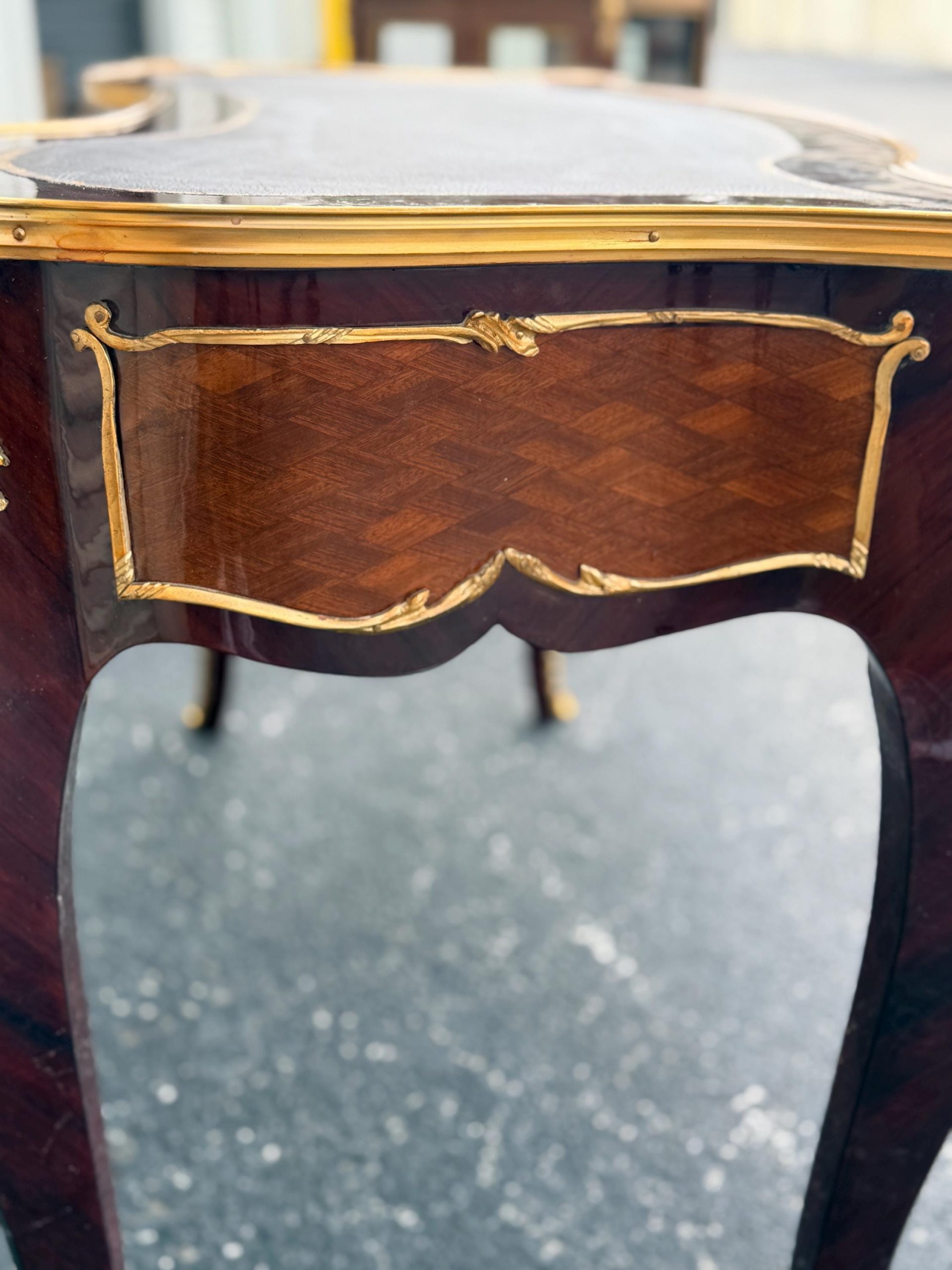 May include: Close-up of a mahogany-colored table with a dark top and gold trim. The table features a decorative inlaid panel with a geometric pattern and ornate gold accents. The legs are curved and the overall design is elegant.