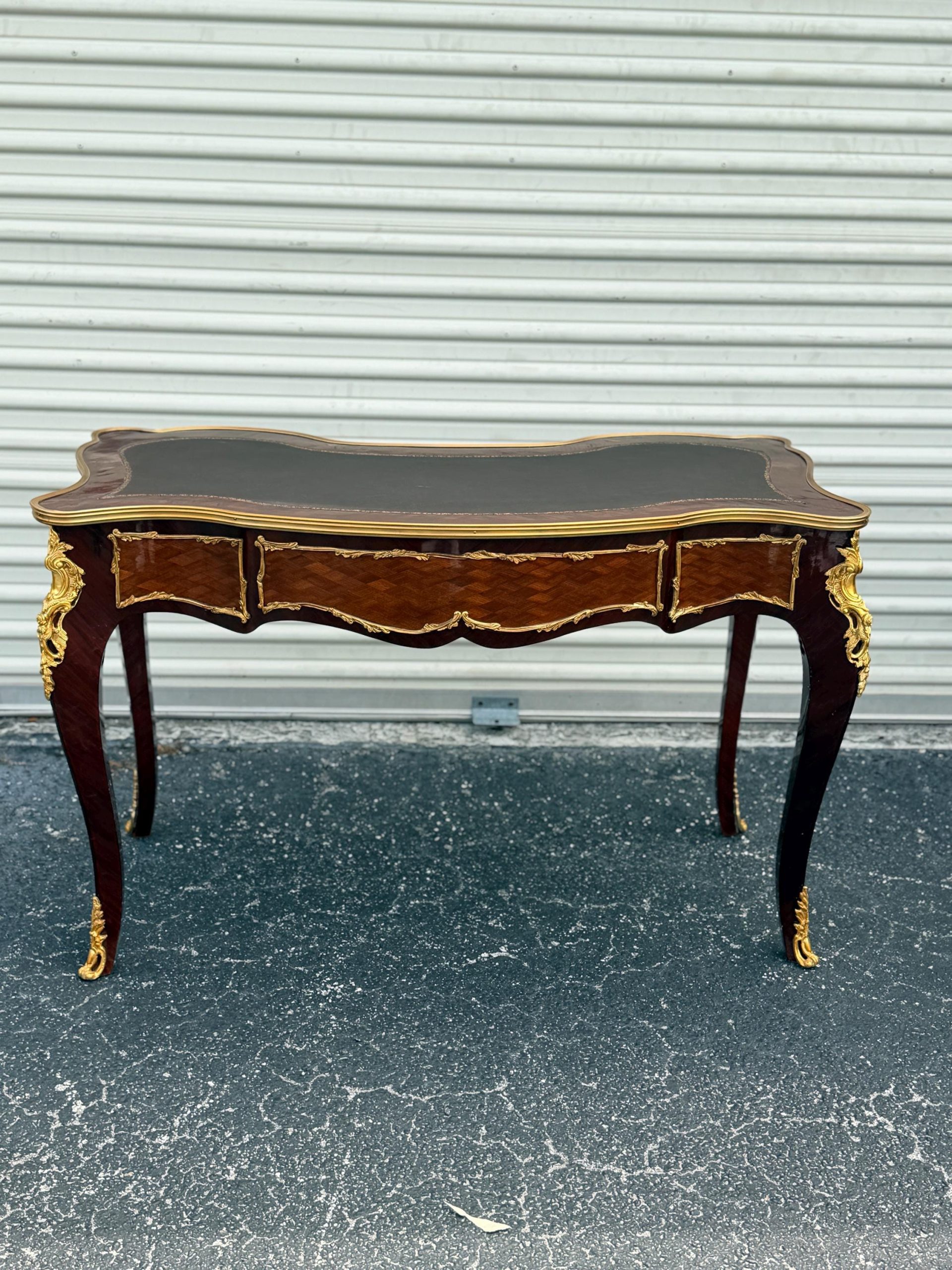 May include: A dark wood desk with a black leather top and three drawers. The desk has ornate gold accents on the legs and edges. The desk is a classic piece of furniture.
