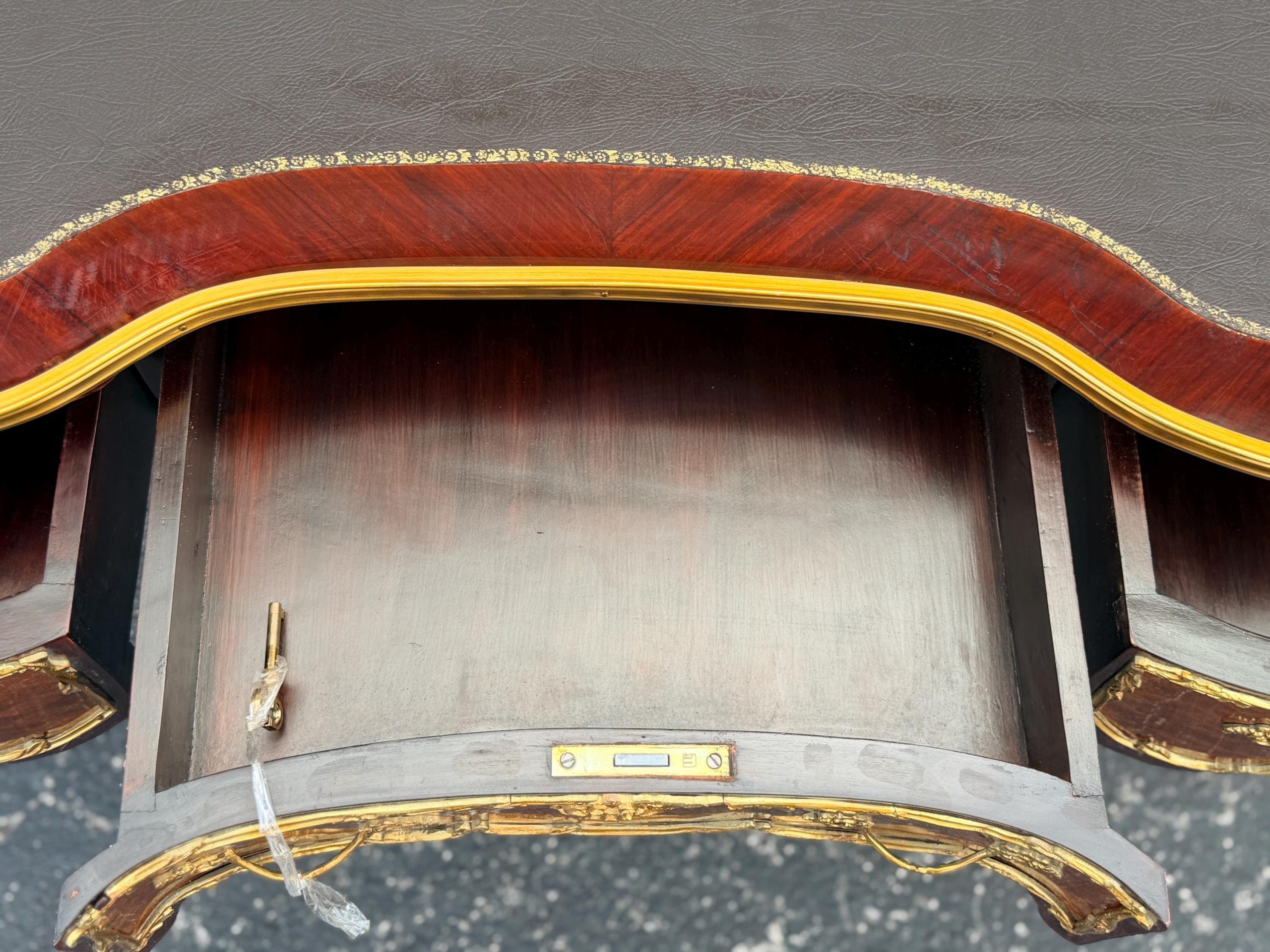 May include: A close-up of a wooden desk with a drawer. The desk has a dark brown top with a reddish-brown border and gold trim. The drawer is open, revealing a light brown interior and a gold-colored lock.