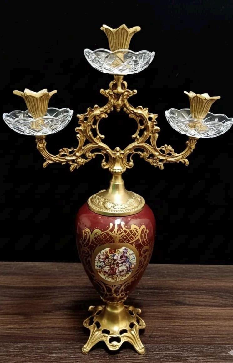 May include: A three-armed candelabra featuring a deep red ceramic base with gold filigree and a central floral medallion. The structure includes ornate gold metal scrollwork, three clear glass candle cups, and a gold metal pedestal base.