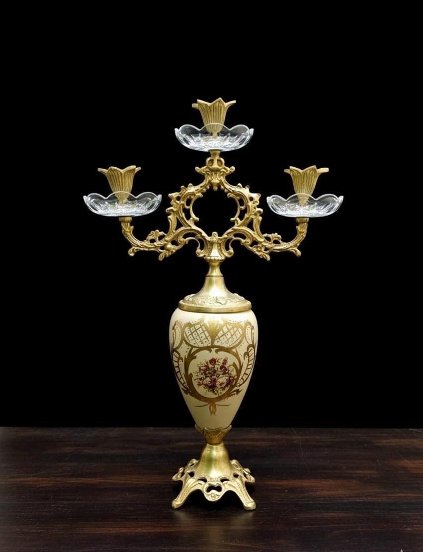 May include: A three-armed candelabra featuring a cream-colored ceramic vase base with gold floral patterns. The top section is made of ornate gold-toned metal with three clear glass candle holders shaped like flower petals at the ends of the arms.