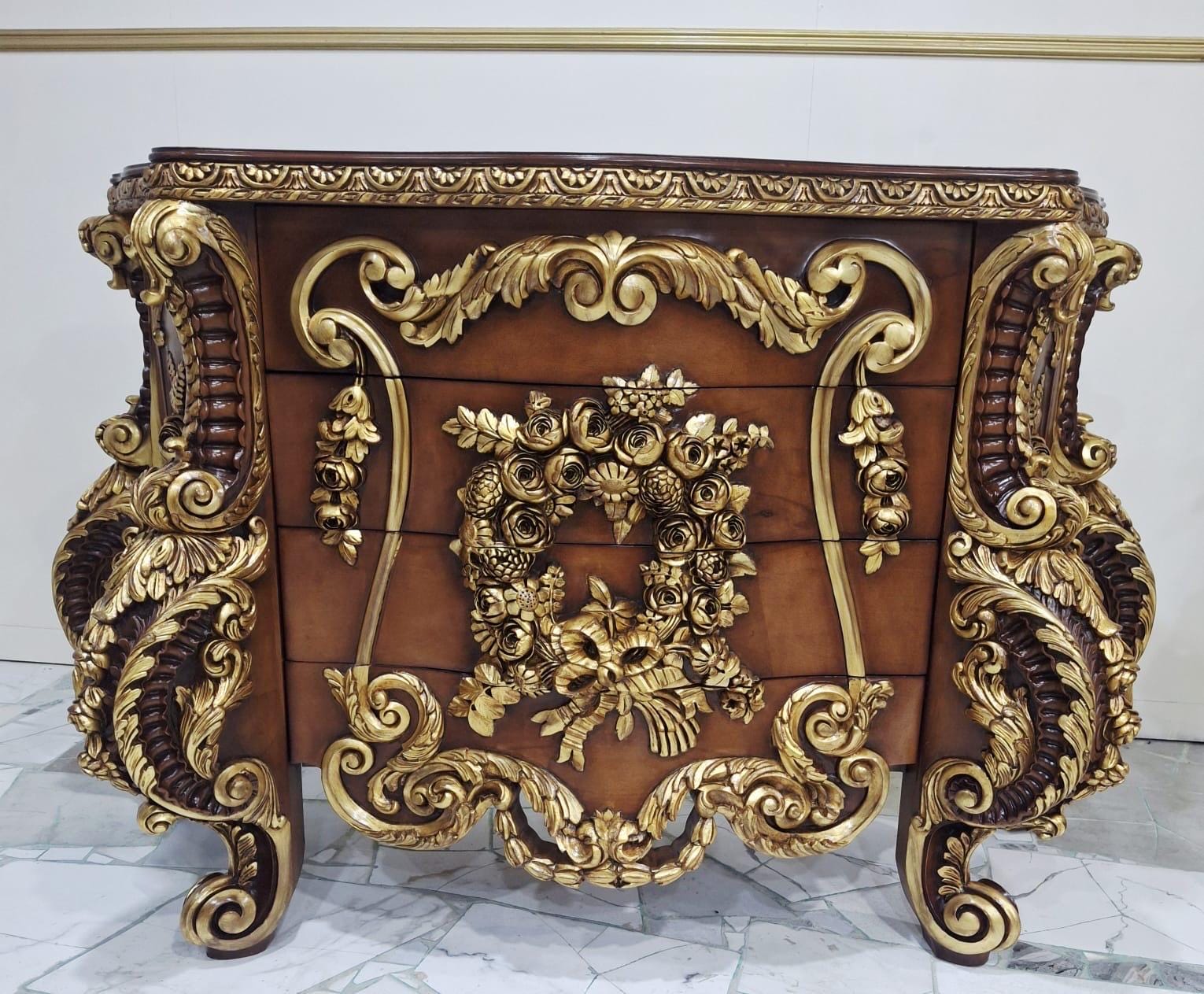 May include: A wooden chest of drawers featuring a rich brown finish and elaborate gold-toned carved ornamentation. The front displays a central floral wreath motif, while the sides and edges are adorned with intricate scrollwork and leaf details.