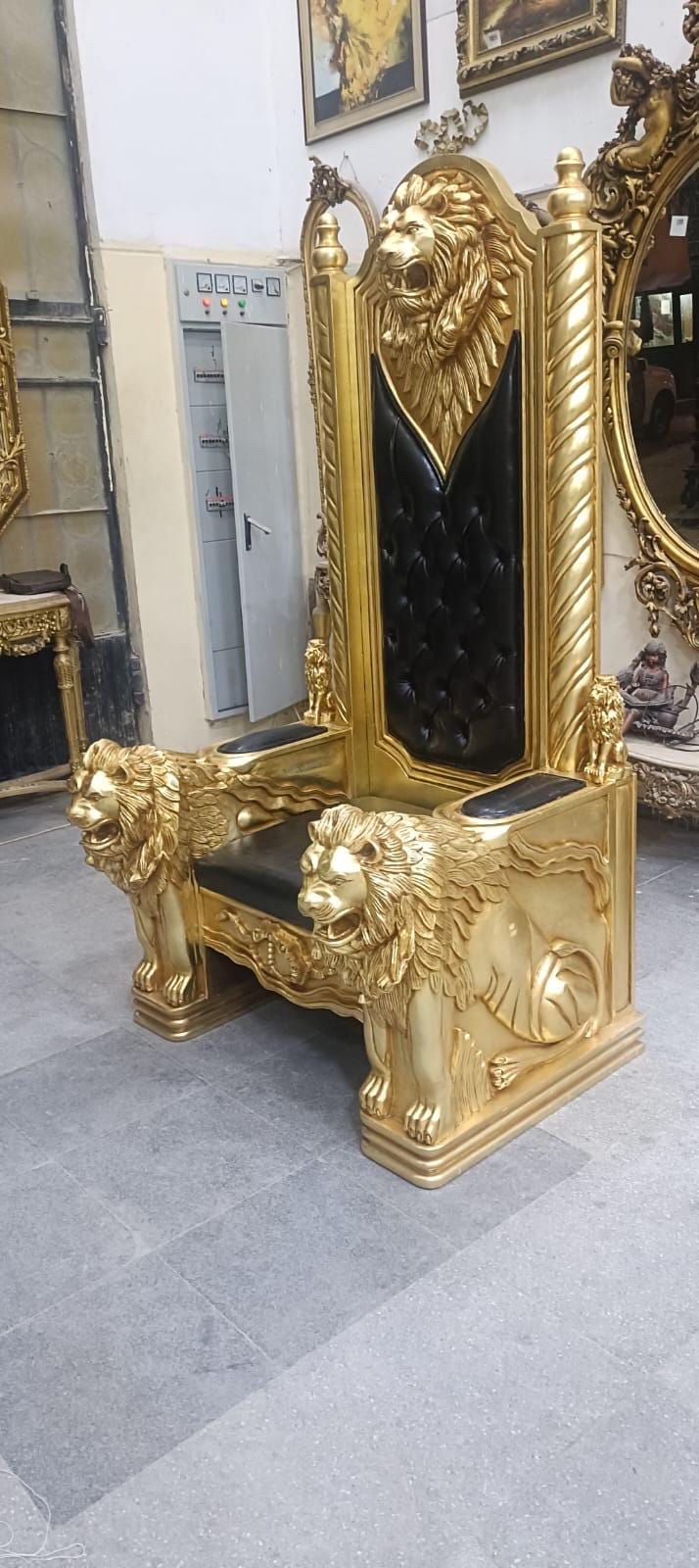 May include: A tall, ornate throne chair featuring a gold-painted wooden frame with detailed lion head carvings on the armrests and the top of the backrest. The seat and backrest are upholstered in black tufted leather, accented by two smaller lion figures.