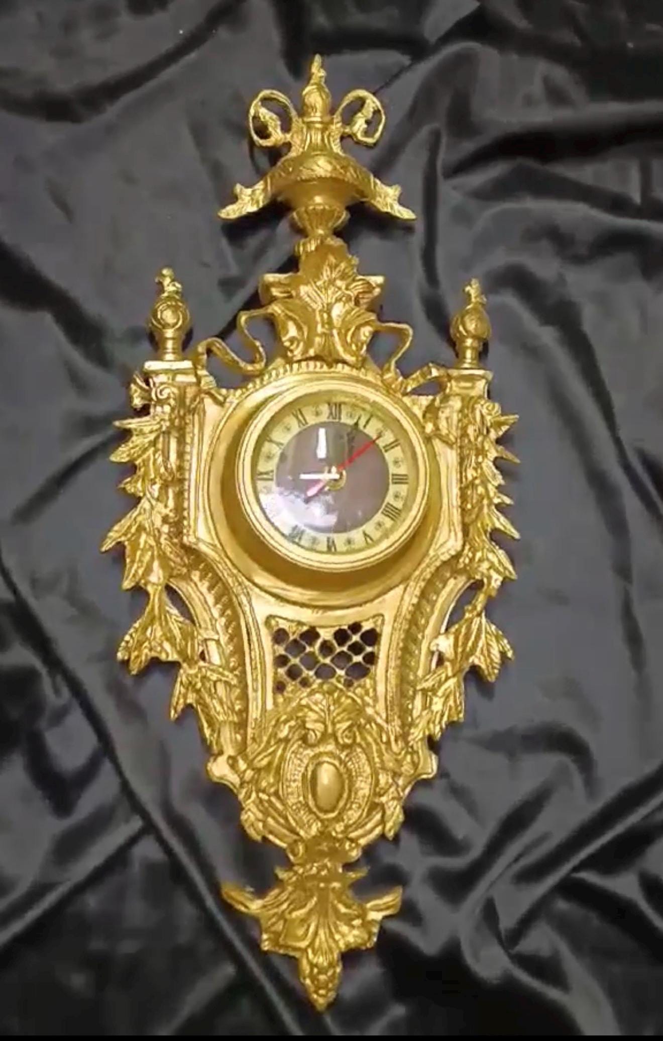 May include: A gold-colored wall clock featuring an ornate, symmetrical frame with intricate leaf and scrollwork detailing. The clock face displays black Roman numerals, a white dial, and three hands, including a red second hand, set within a decorative casing.