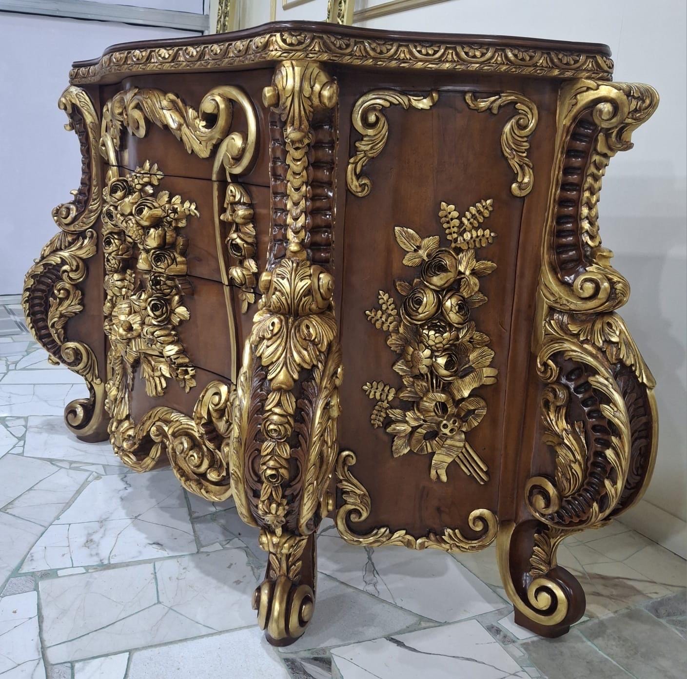 May include: A wooden chest of drawers featuring a dark brown finish with ornate gold-colored carved accents. The piece displays intricate floral relief carvings on the front and sides, curved legs, and a decorative border along the top edge.