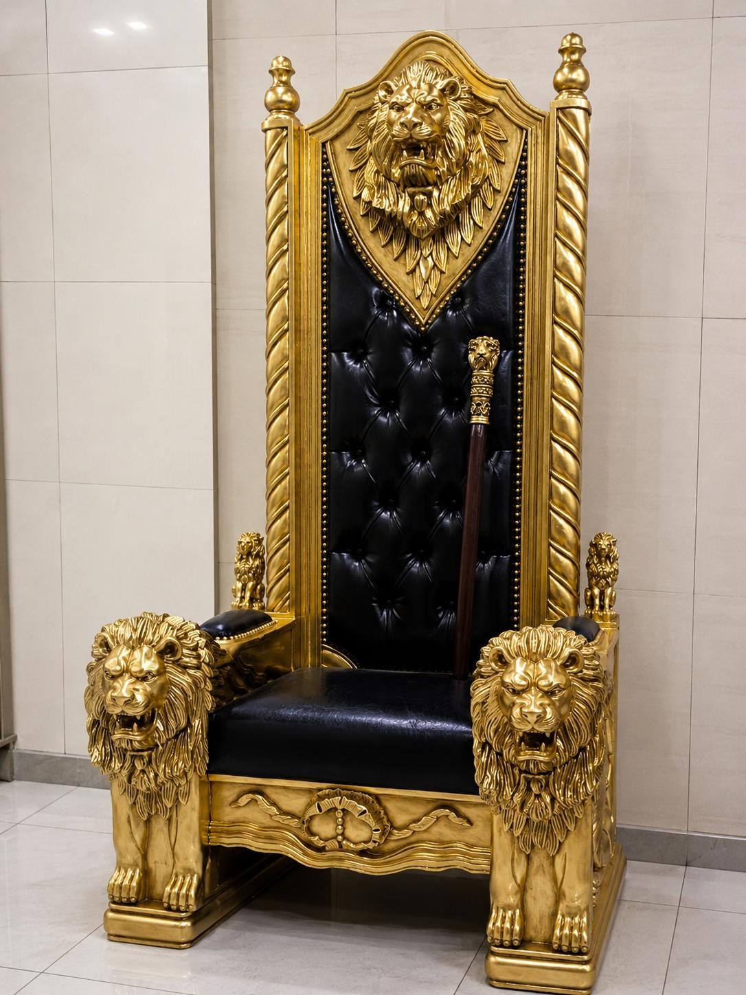 May include: A tall, ornate throne featuring a black tufted leather seat and backrest. The frame is finished in gold, with detailed lion head carvings on the armrests and the top center. A wooden staff with a gold lion head topper rests on the seat.