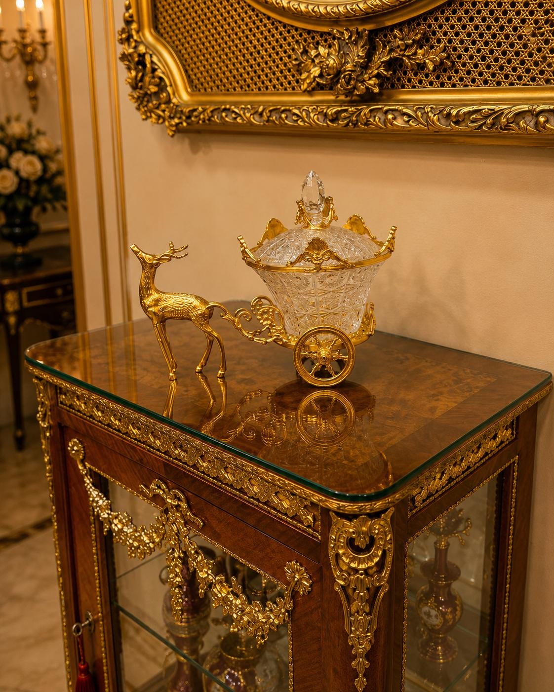 May include: A decorative centerpiece featuring a clear, faceted glass bowl with a lid, shaped like a carriage pulled by a gold-toned metal deer figure. The carriage has gold-toned wheels and ornate metal detailing, resting on a glass-topped wooden cabinet.