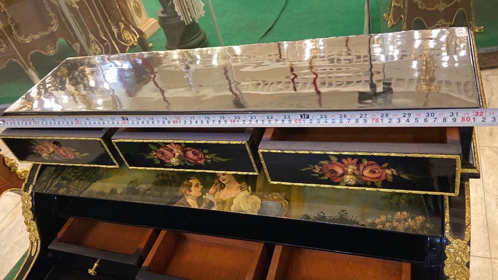 May include: A black lacquered desk with gold trim and floral painted drawer fronts. A measuring tape across the top shows a width of approximately 32 inches. The desk features multiple drawers and a painted scene of two figures on the interior surface.