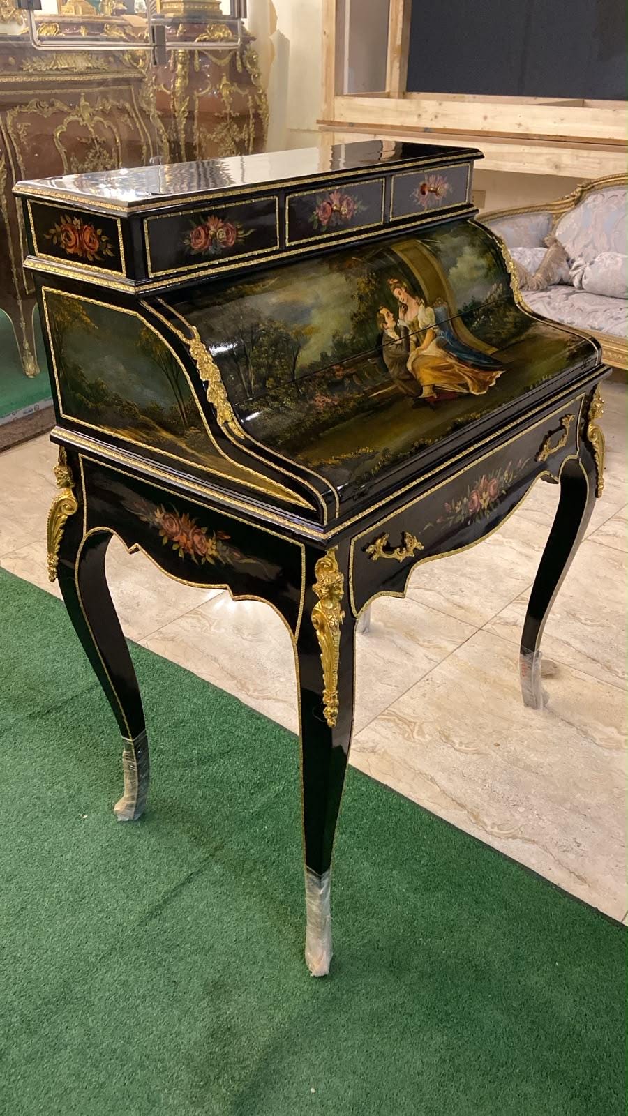 May include: A black lacquered roll-top desk featuring gold trim, ornate floral hand-painted accents, and a detailed scene of two figures in a landscape on the curved lid. The desk stands on four slender legs with gold-toned metal decorative mounts.