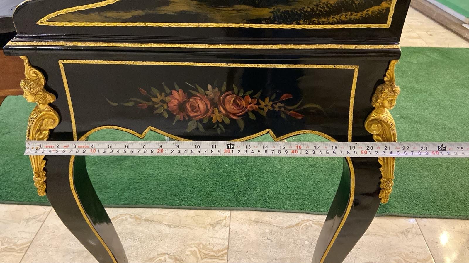 May include: A black lacquered console table with gold trim, featuring a hand-painted floral motif of roses on the front apron and ornate gold-toned metal mounts on the legs. A measuring tape shows the table width is approximately 24 inches.