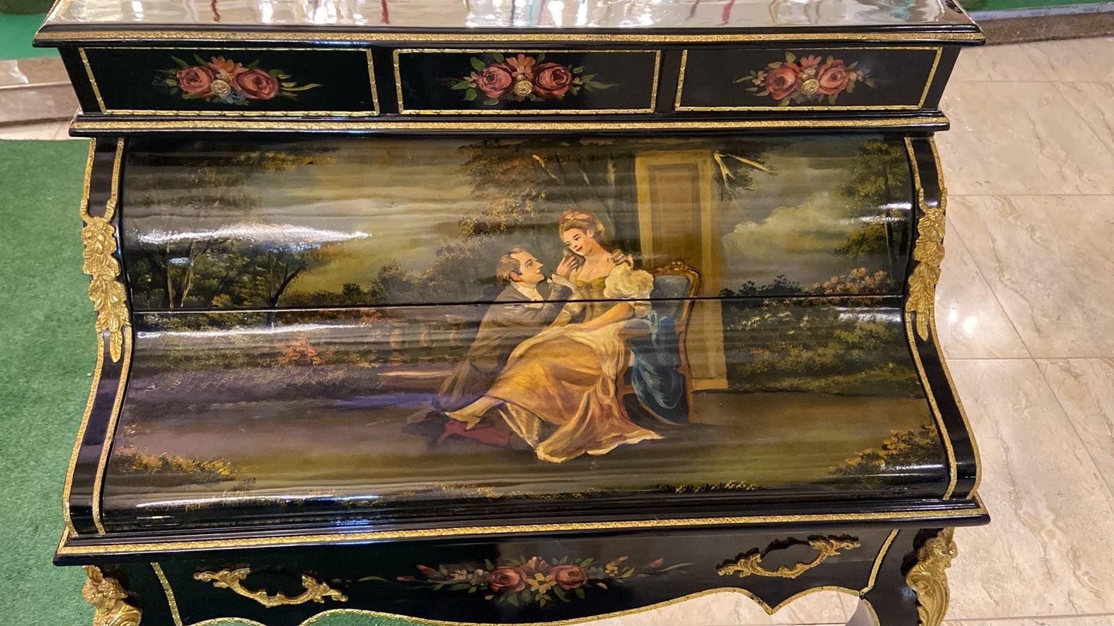 May include: A black lacquered roll-top desk featuring gold-toned metal accents and ornate floral motifs on the top drawers. The curved front panel displays a detailed hand-painted scene of two figures in a garden setting with lush greenery.