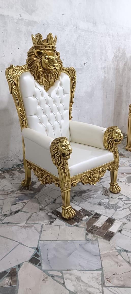 May include: A high-back throne chair featuring a gold-painted wooden frame with intricate lion head carvings at the top and on the armrests. The backrest is upholstered in white tufted leather with button accents, and the legs end in lion paw feet.