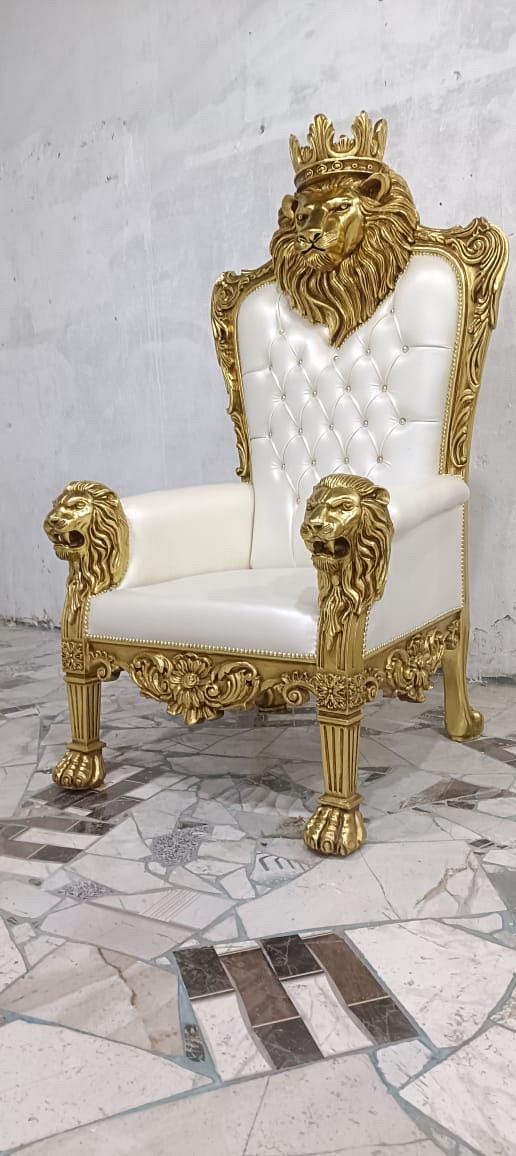 May include: A high-backed throne chair featuring a white tufted leatherette seat and backrest. The frame is finished in metallic gold, showcasing intricate lion head carvings on the armrests and a large lion head with a crown at the top.