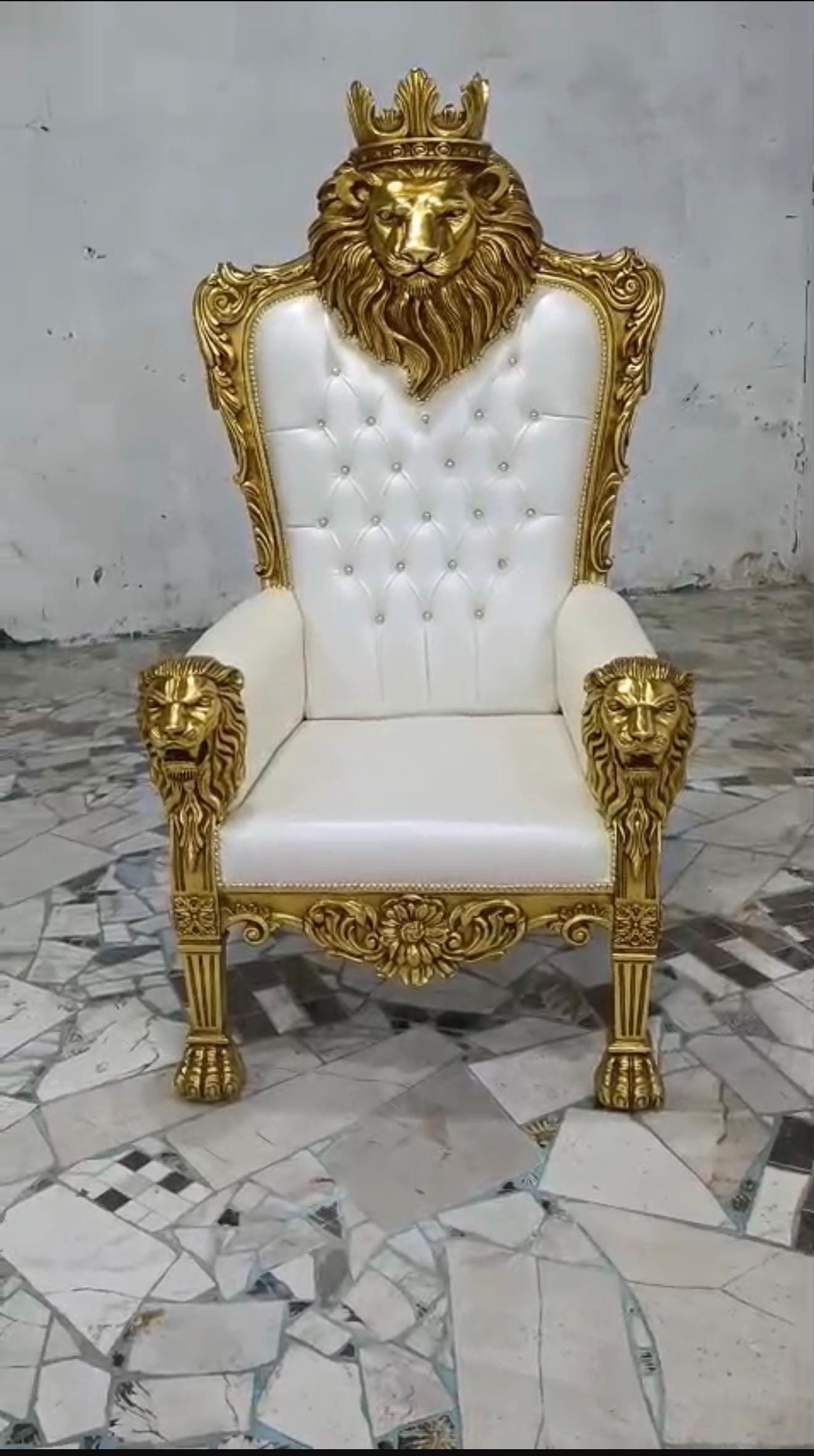 May include: A high-back throne chair featuring a white tufted upholstered seat and backrest. The frame is finished in gold, showcasing a prominent lion head carving at the top center with a crown, and lion head motifs on each armrest.