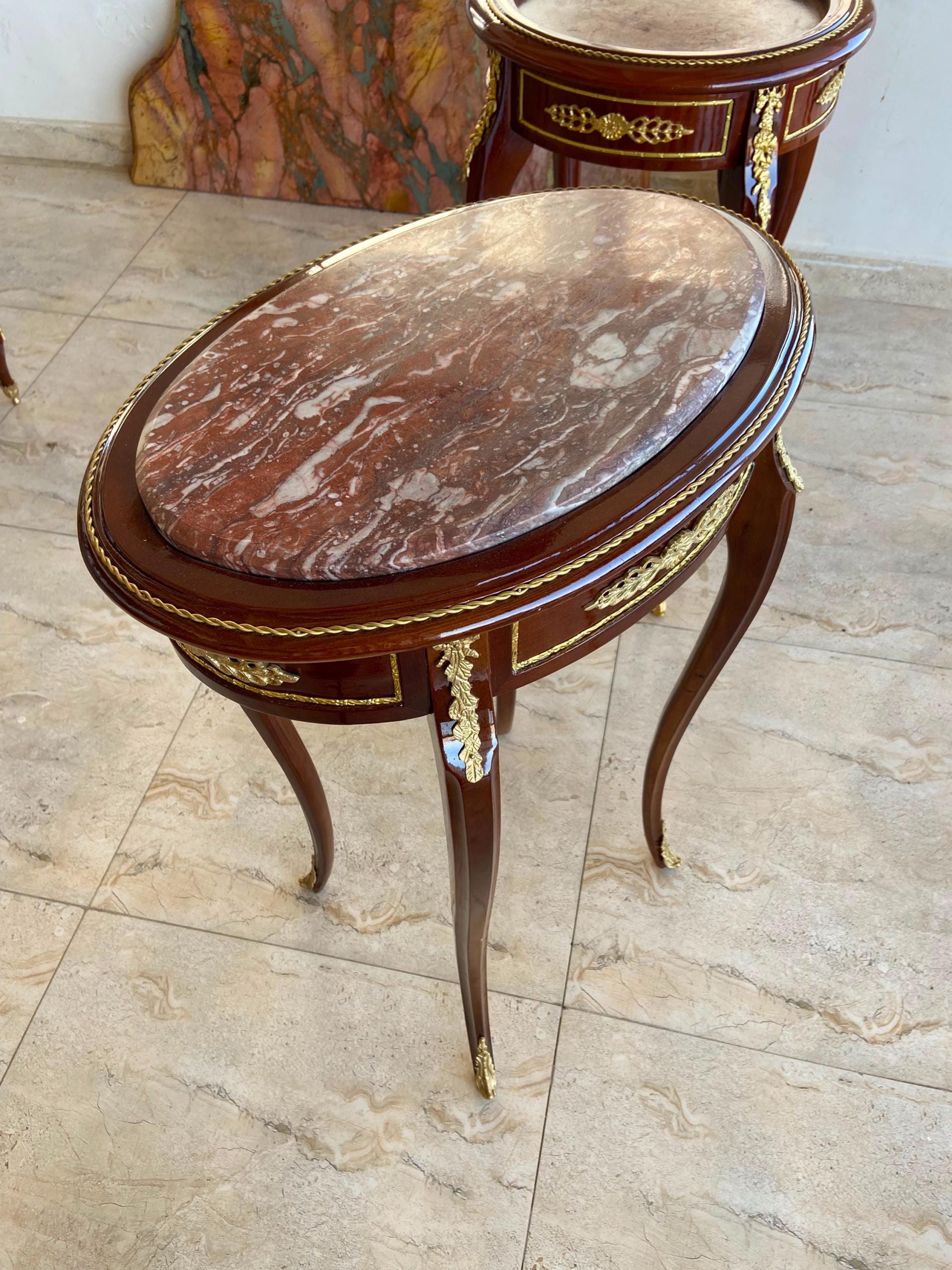 May include: An oval side table with a polished dark wood frame and a red and white marbled stone top. The table features curved legs and gold-toned metal decorative accents along the apron and legs. A second matching table is visible behind it.