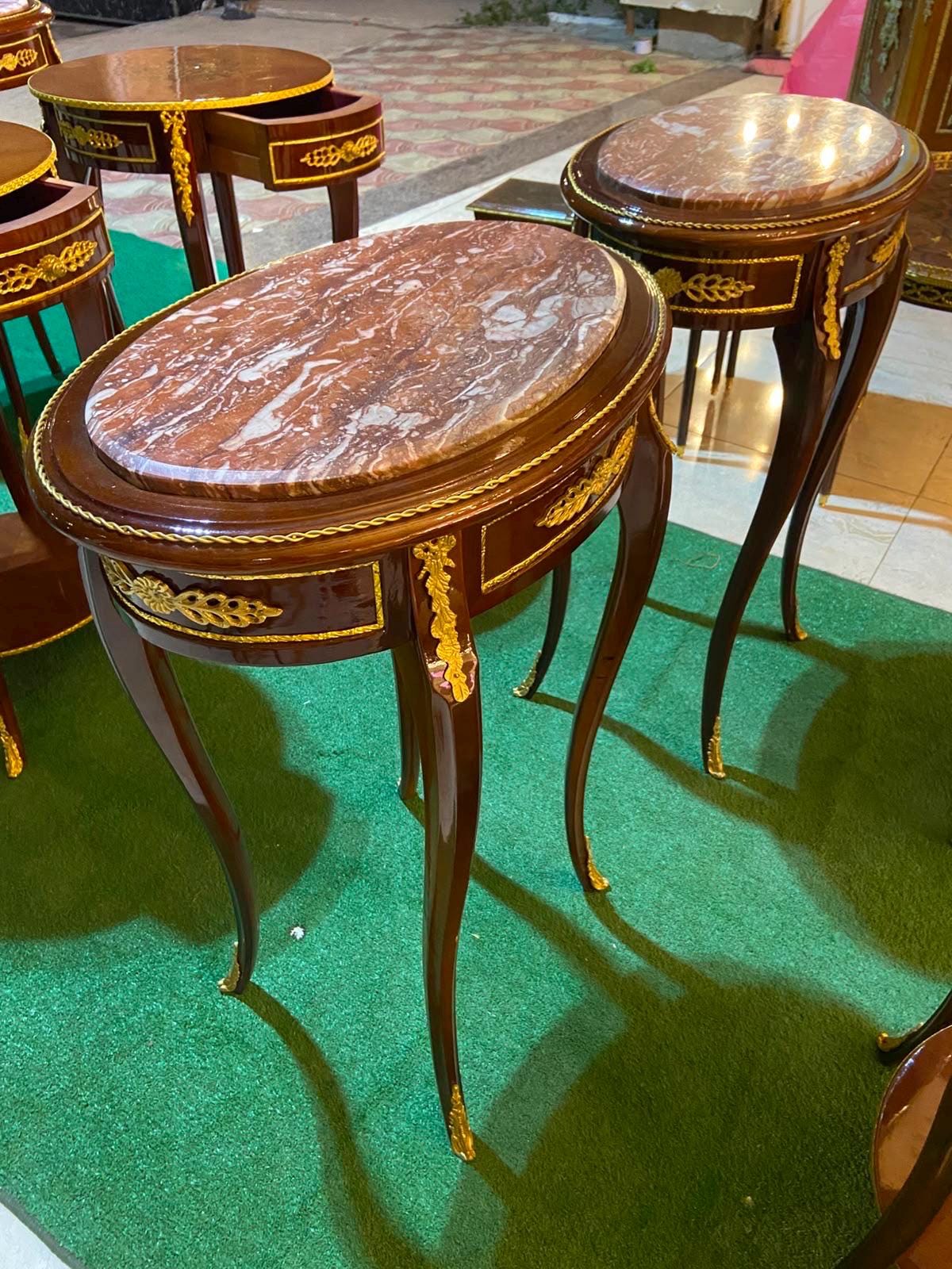 May include: A set of oval side tables with dark brown wooden frames and polished marble tops. Each table features a single drawer with gold-colored metal leaf ornamentation, a rope-style trim around the edge, and four slender, curved legs.