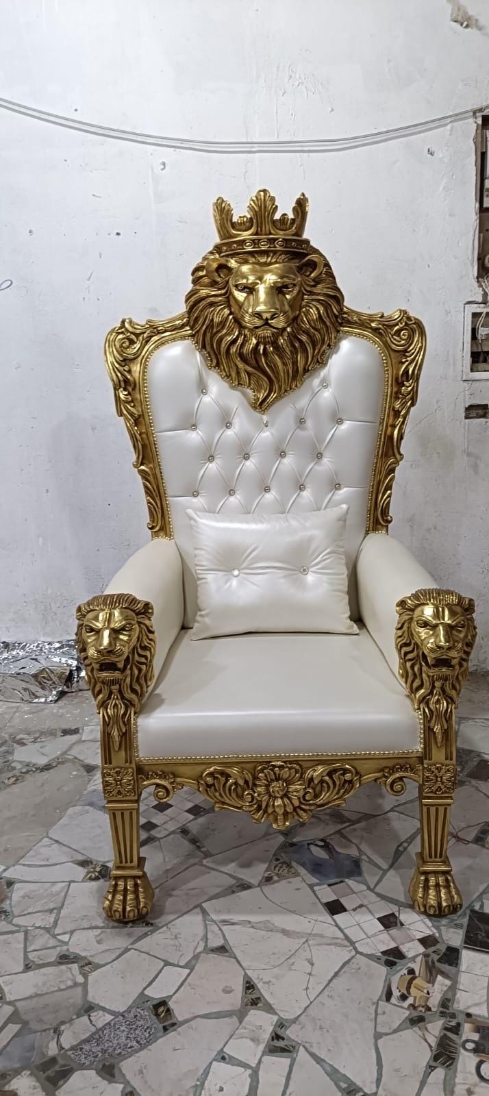 May include: A high-back throne chair featuring a gold-painted wooden frame with carved lion heads on the armrests and a large lion head with a crown at the top. The seat and back are upholstered in white tufted leather with a matching white throw pillow.