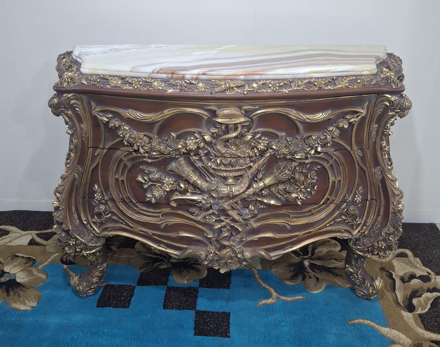 May include: A wooden chest of drawers featuring a polished marble top with brown and white veining. The front is adorned with intricate gold-toned carvings, including floral motifs and cornucopia designs, set against a dark brown wood finish.