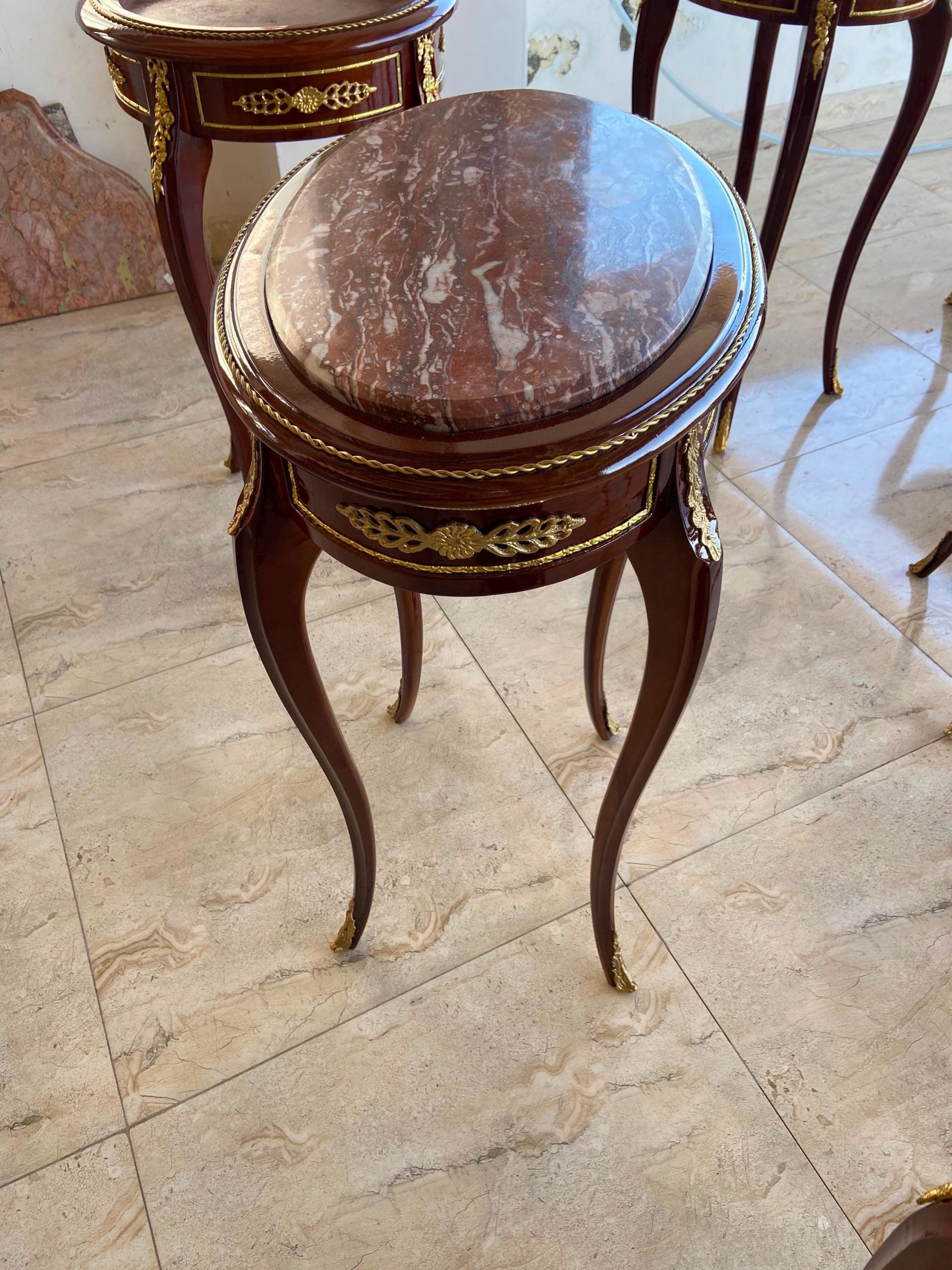 May include: An oval side table with a polished dark wood frame and a reddish-brown marble top. The table features curved legs, gold-toned metal decorative accents, and a rope-style trim around the edge of the tabletop surface.