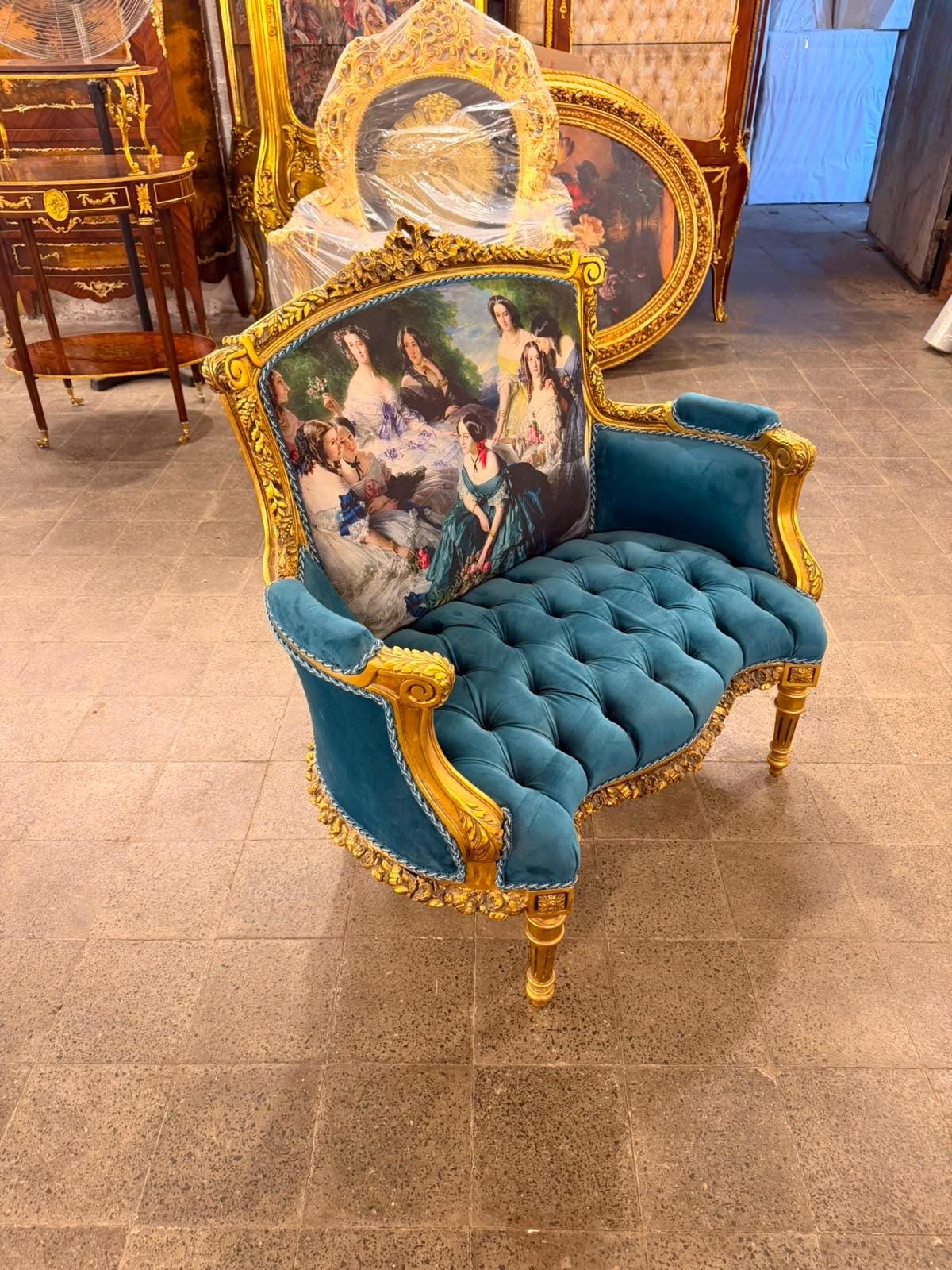 May include: An armchair with a gold-painted wooden frame featuring intricate carvings. The backrest displays a printed scene of several figures in period clothing. The seat and armrests are upholstered in teal velvet with button tufting.