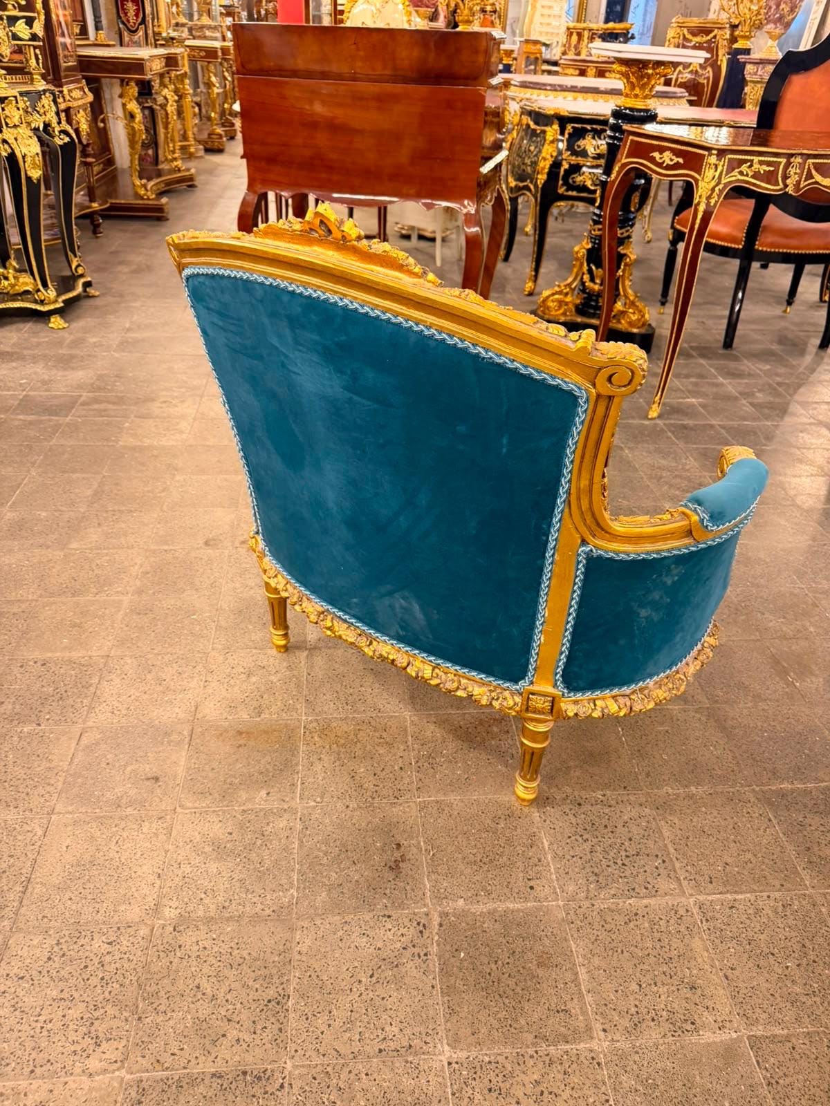 May include: A teal velvet settee featuring a gold-painted wooden frame with intricate carved floral details along the base and back. The upholstery is accented with a light blue twisted cord trim along the edges, supported by four tapered legs.