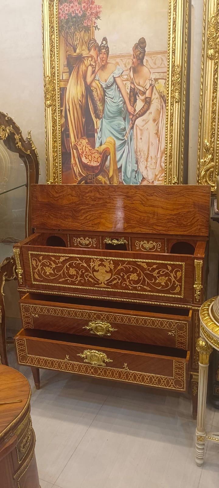 May include: A wooden chest of drawers featuring a rich brown wood grain finish, gold-toned metal hardware, and intricate gold floral inlay patterns. The piece includes a top hinged compartment and two lower drawers with ornate brass handles.