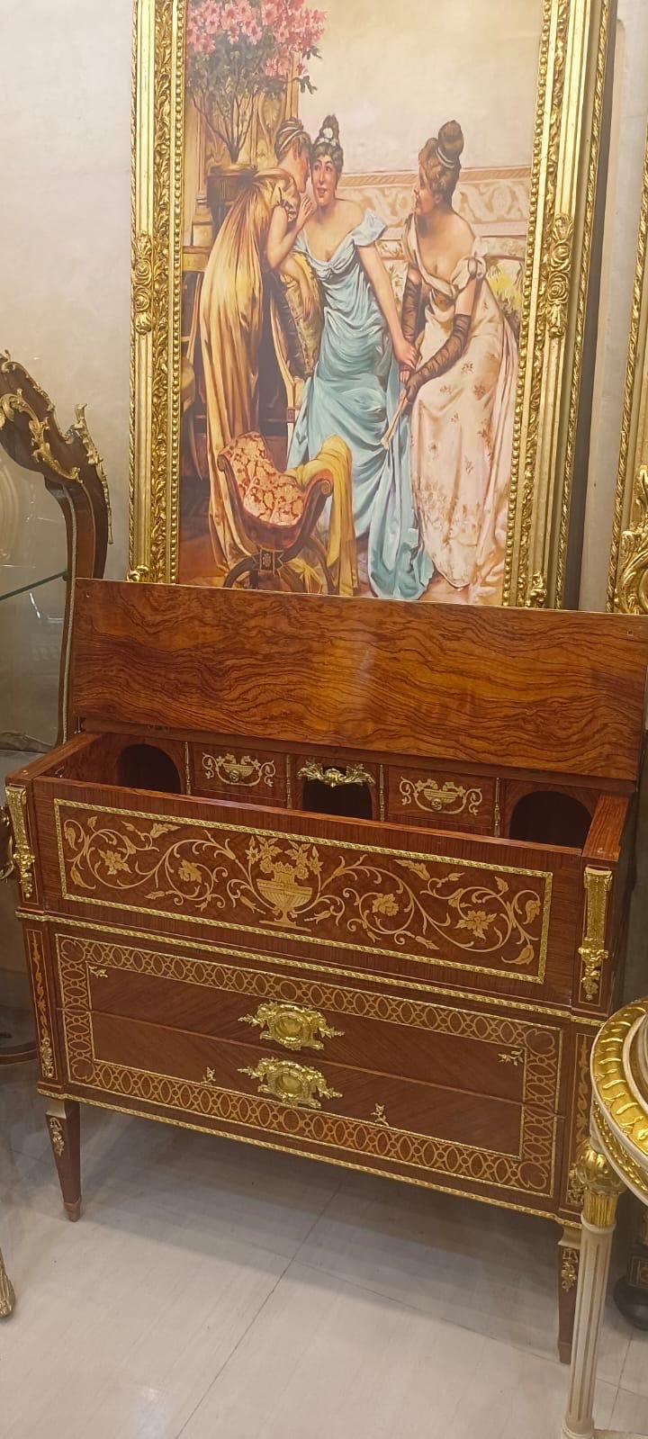May include: A wooden chest of drawers featuring a drop-front desk compartment with an open top. The piece is decorated with intricate gold-toned metal inlays, ornate brass drawer pulls, and gold trim along the edges of the wood grain panels.