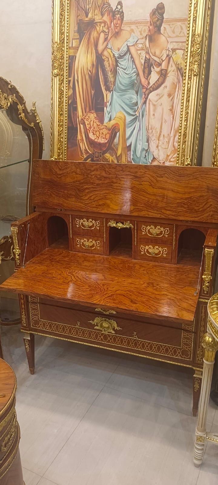 May include: A wooden secretary desk with a drop-down writing surface, featuring a rich wood grain finish, gold-toned metal hardware, and ornate decorative accents. The desk includes multiple small drawers and open compartments for storage.