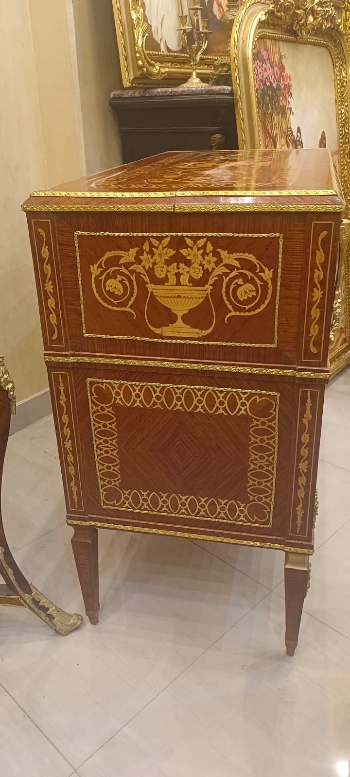 May include: A rectangular wooden cabinet featuring dark brown wood veneer with intricate gold-toned metal inlay and trim. The upper panel displays a central gold floral urn motif, while the lower panel showcases a geometric border pattern.