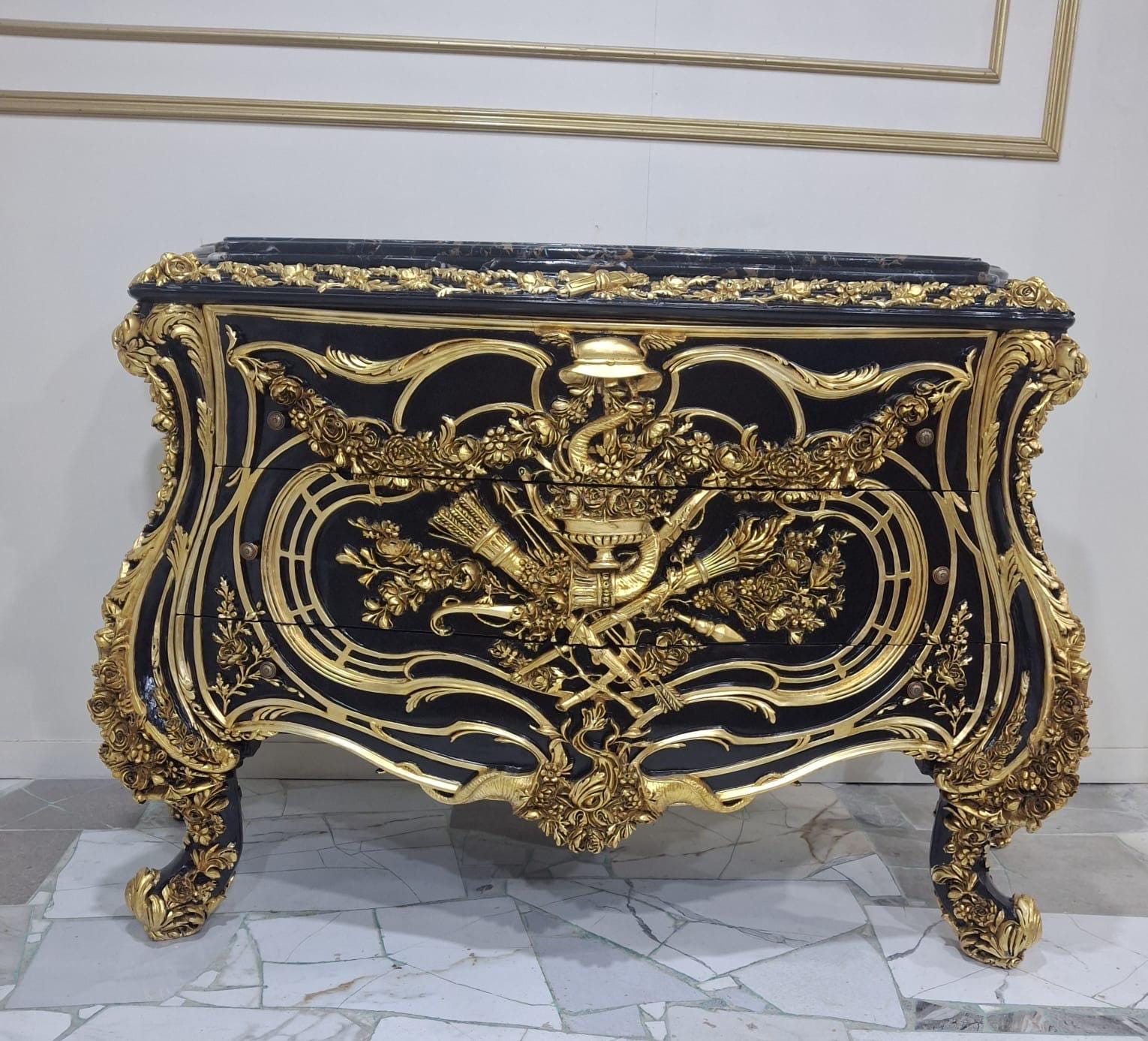 May include: A black chest of drawers featuring ornate gold-colored metal ornamentation. The front displays intricate relief carvings of floral motifs, a central horn, and arrows. The piece has curved legs and a dark marble top with gold trim.