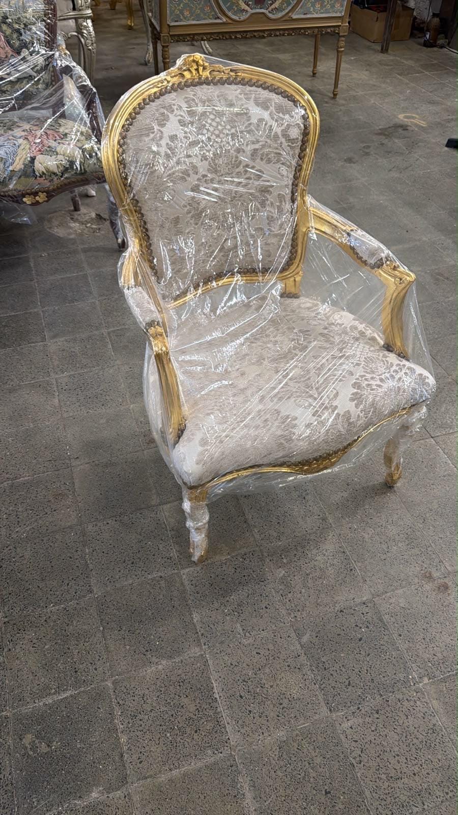 May include: An armchair with a gold-painted wooden frame and a high, rounded backrest. The seat and back are upholstered in a cream-colored fabric featuring a subtle, repeating floral damask pattern. The entire chair is wrapped in clear plastic.
