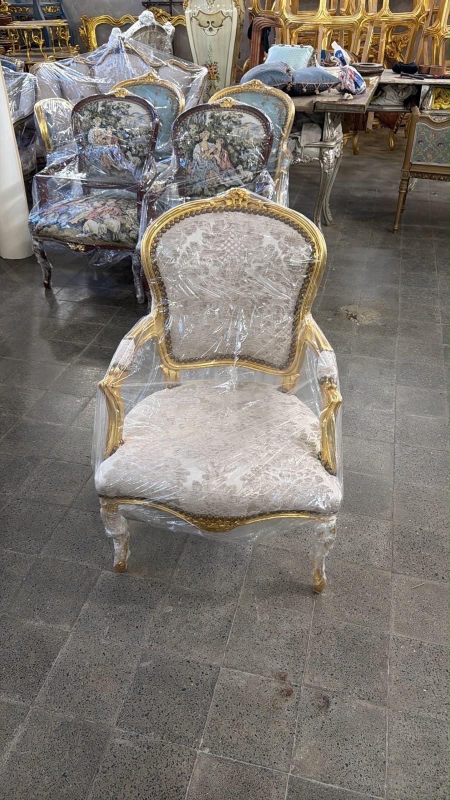 May include: An armchair with a gold-painted wooden frame and light-colored fabric upholstery featuring a subtle damask pattern. The chair has curved armrests, a rounded backrest, and cabriole legs, all wrapped in clear protective plastic film.