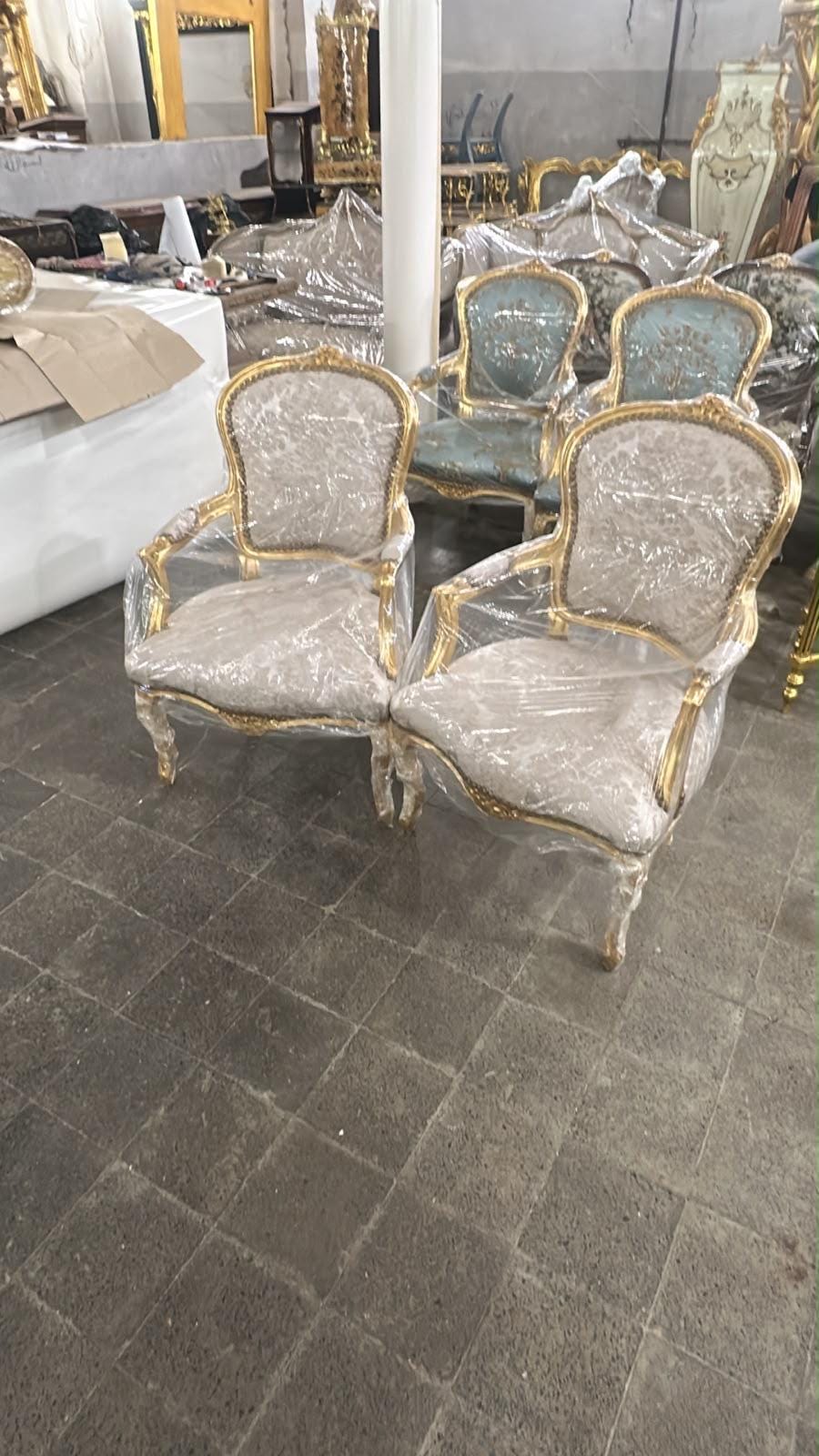 May include: Two armchairs with gold-painted wooden frames and light-colored patterned fabric upholstery. Each chair features curved backrests, padded armrests, and cabriole legs. The chairs are wrapped in clear protective plastic film for storage.