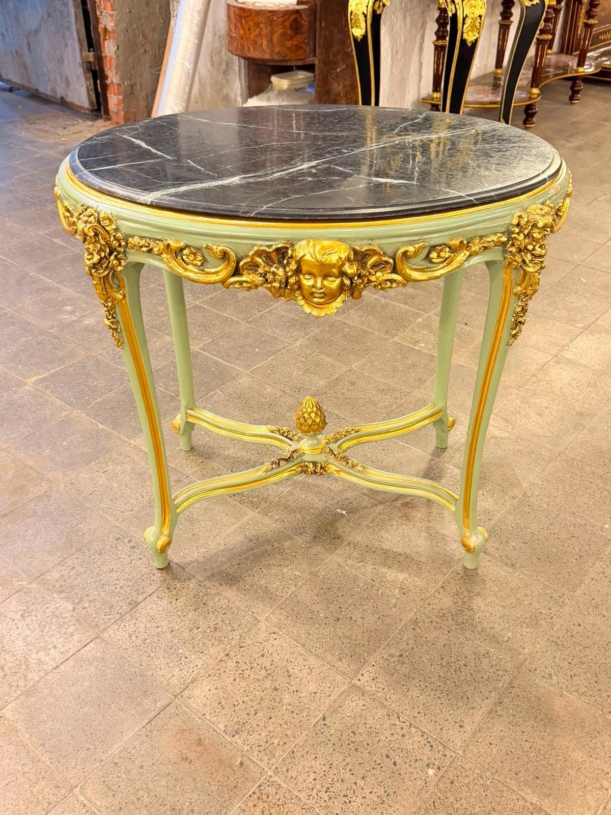 May include: A round side table with a black marble top and a light green frame. The table features ornate gold-colored carvings, including a central cherub face on the apron, floral motifs on the legs, and a decorative finial at the center.